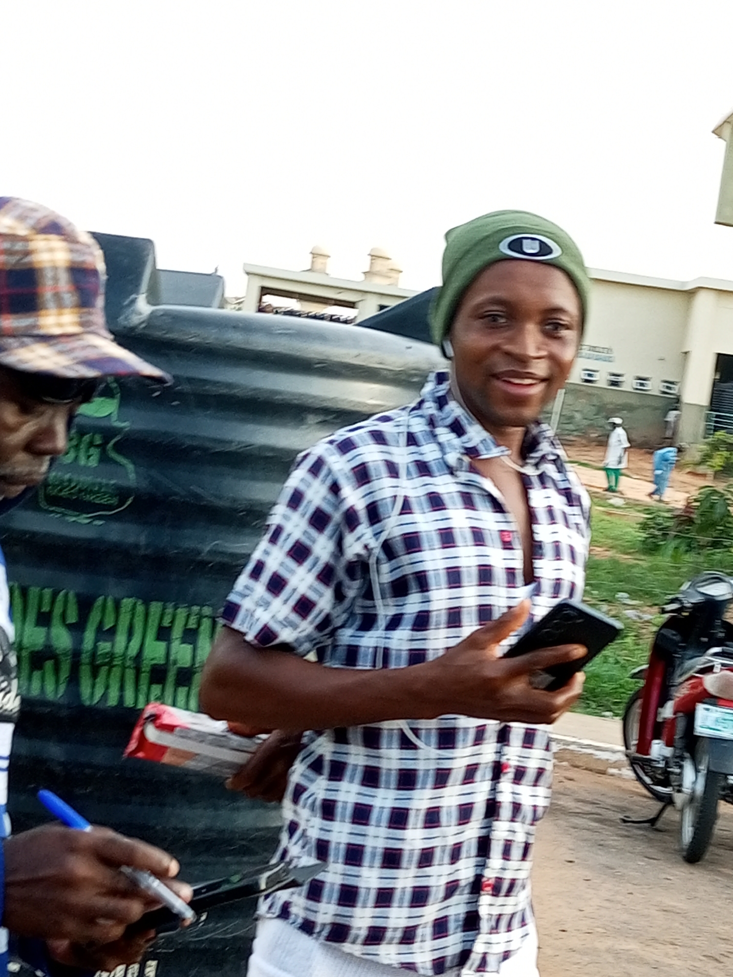 In Gombe, Nigeria, a man wearing a green beanie and a plaid shirt is holding a smartphone and looking at the camera. In the background, a motorcycle is parked, and two other individuals are visible. The scene appears to be outdoors, possibly on a street or in a public area, with buildings and vegetation present. The man in the foreground is the primary subject, and his engagement with the smartphone suggests he may be taking a picture or engaging with an application.