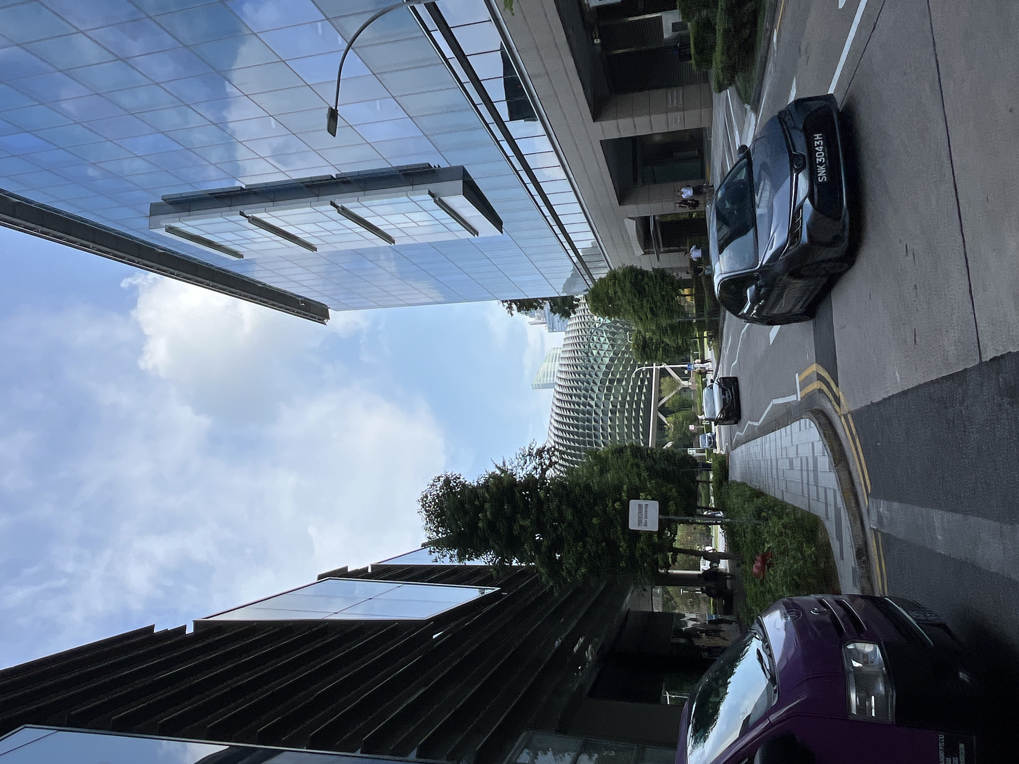 The image depicts a street scene in Singapore, with modern glass-fronted buildings on both sides of a road. In the background, the distinctive durian-shaped roof of the Esplanade – Theatres on the Bay is visible. A dark gray sedan with license plate SNK 3043H is driving on the road, and a portion of a purple van is visible in the foreground on the left. A sidewalk with trees and a pedestrian sign runs alongside the buildings. The sky is partly cloudy.