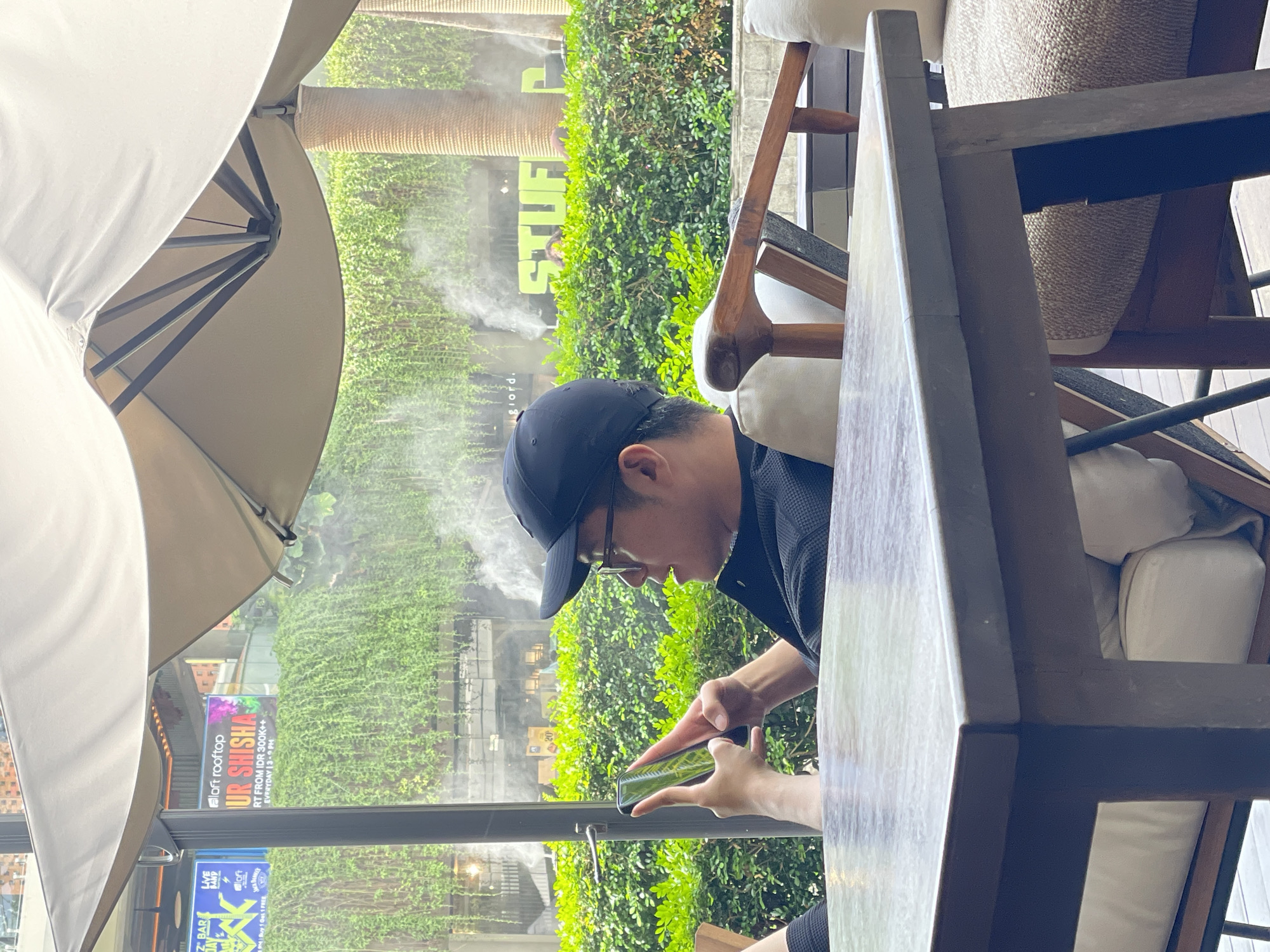 A young Asian male wearing a black baseball cap and glasses is seated at an outdoor table, looking at his mobile phone. He is wearing a black collared shirt. The table is made of wood with a textured, greyish-brown surface. In the background, there is lush green foliage, misting sprayers, and a building with signage that appears to be in English. One sign reads "loft rooftop YOUR SHISHA" and another sign has yellow text that includes "STUFF". The scene suggests an outdoor dining or lounging area in a warm climate. The location is Kuta, Indonesia.