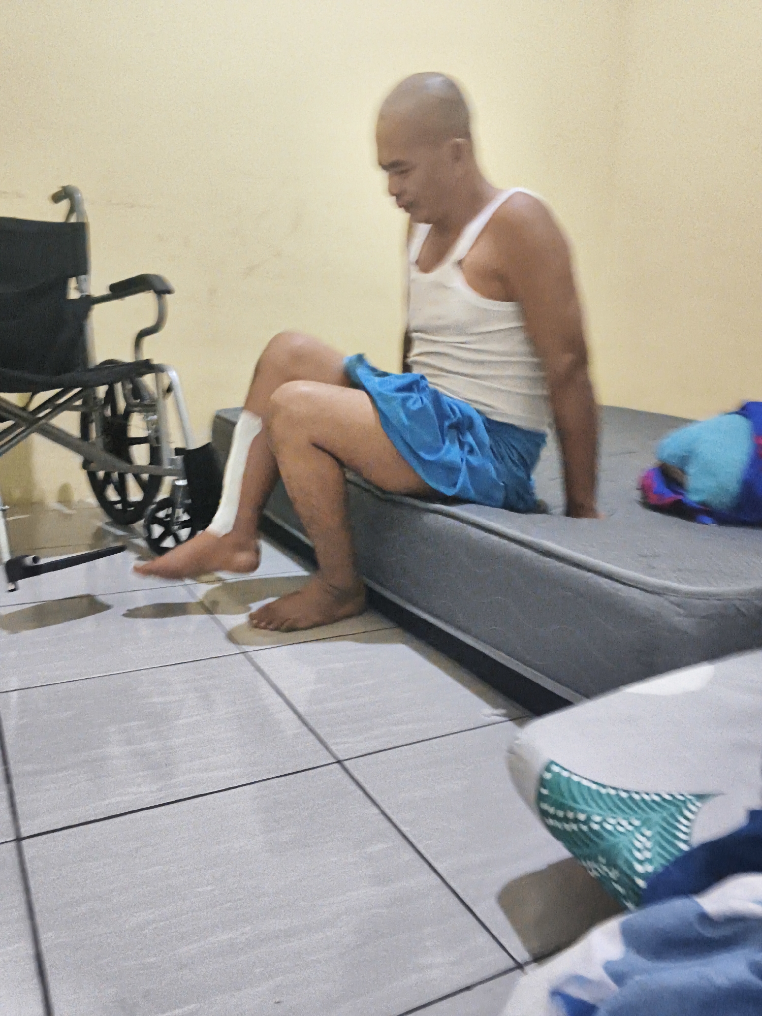 The image depicts a dimly lit indoor scene, likely a room in Banjar Agung, Indonesia. A bald man wearing a white tank top and blue shorts is seated on a grey mattress. His left leg is bandaged from the ankle up to just below the knee. Next to the mattress, to the left of the man, is a black wheelchair. The floor is tiled with grey square tiles. In the background, a pale yellow wall is visible. A colorful blanket or comforter is on the mattress, to the right of the man.