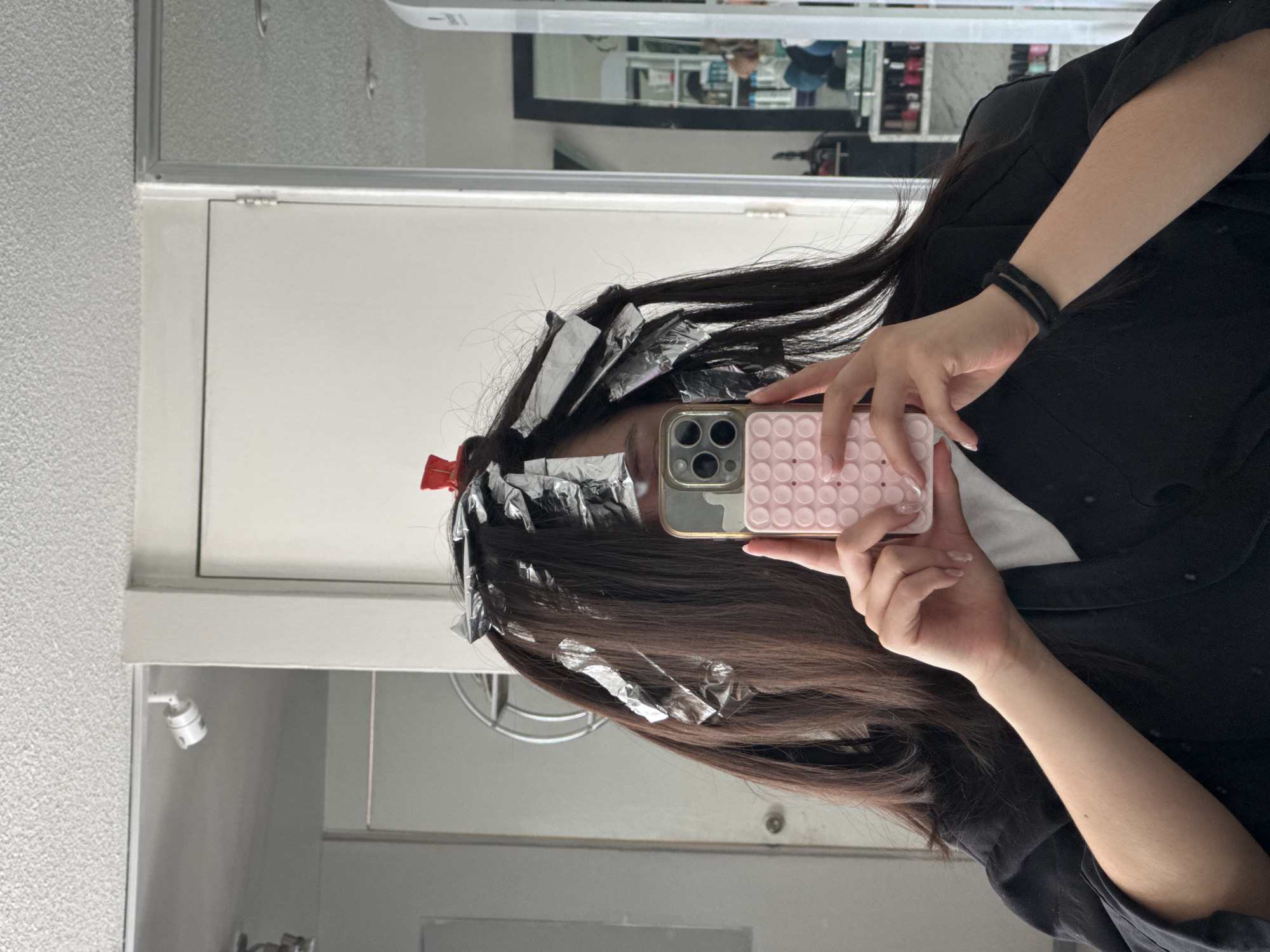 A person is taking a selfie in what appears to be a salon or bathroom setting. The person is having their hair highlighted or dyed, as evidenced by the foil applied to sections of their hair. They are holding a pink and gold iPhone in front of their face. The person is wearing a black top and a black bracelet. The background includes a white wall, a mirror, and what looks like a cosmetics display in the reflection of the mirror. No evidence suggests the city of Puebla, Mexico.