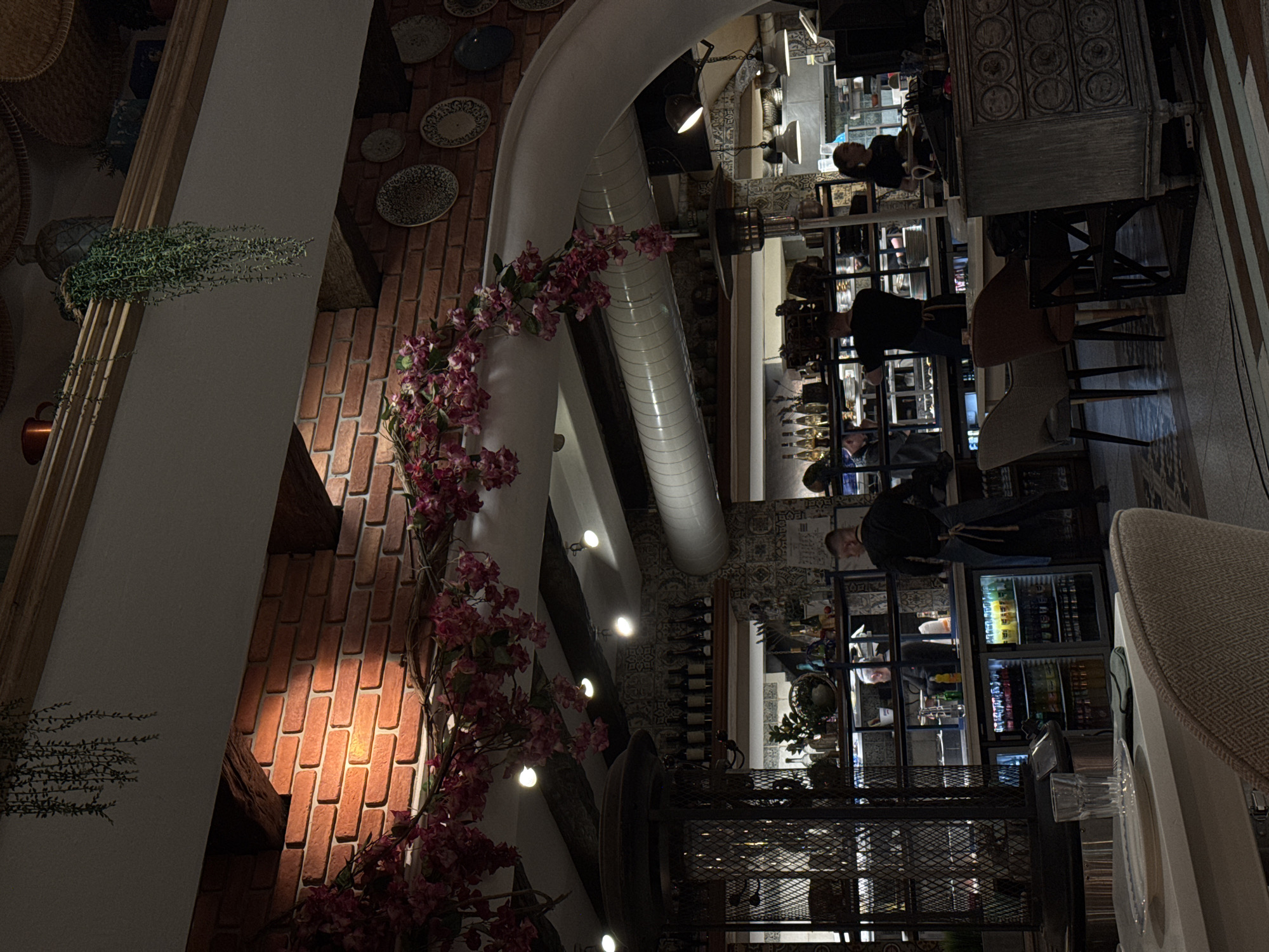 A distorted overhead view captures the interior of a restaurant, likely in Varna, Bulgaria, blending architectural elements with human activity.
The foreground features a brick wall adorned with decorative plates and lit by recessed lighting, contrasting with a white arched structure embellished with pink floral arrangements. A metallic ventilation duct is visible behind the arch.
The central and background areas depict restaurant staff behind a serving counter stocked with beverages and food items. Patrons are present, one visible behind the counter. Tables and chairs are scattered throughout the scene, suggesting a dining area.
The environment includes architectural details such as a decorative tiled floor and a metal shelving unit, contributing to the restaurant's ambiance.