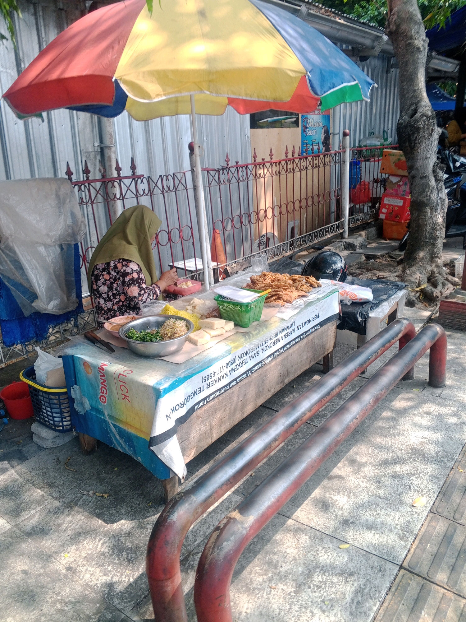 This image depicts an outdoor food stall in Wonogiri, Indonesia, under a colorful umbrella offering shade. A woman wearing a hijab and a floral-patterned shirt is preparing food. On the table, various ingredients are visible, including a bowl of sprouts and noodles, blocks of tofu, and a plate of fried food. The stall is set up on a sidewalk, with metal railings in the foreground and a corrugated metal wall in the background. Boxes are stacked near a tree to the right. The overall scene suggests a daytime setting, likely morning or afternoon, given the sunlight casting shadows. The text visible on the tablecloth includes a warning about smoking and a contact number for a smoking cessation service in Indonesian. The presence of a tree and the architecture suggest a typical urban or semi-urban environment in Indonesia.