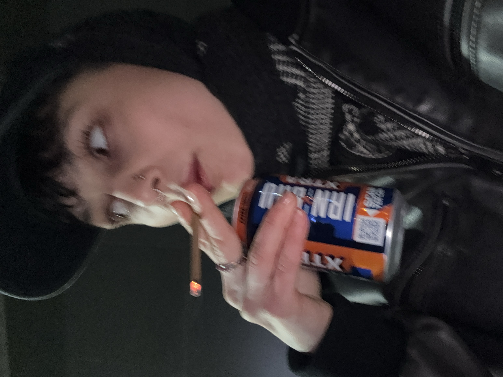The original image is rotated 90 degrees counter-clockwise, capturing a close-up of a person, likely a young adult, with dark hair and bangs. They have a nose piercing and are wearing dark clothing, possibly a black jacket or hoodie.
The individual is holding a lit cigarette or joint in one hand, with a visible ember glowing at its tip. In their other hand, they grasp a can of "IRN-BRU XTRA," featuring its distinct orange and blue design. The can prominently displays the brand name, along with "SCAN & WIN" text and a QR code. Rings are visible on their fingers.
The person's gaze is directed upwards and to their right, conveying a relaxed or contemplative expression. The background is predominantly dark and indistinct, indicating a dimly lit environment, possibly indoors or an outdoor setting at night. No specific geographical or architectural cues are visible within the image frame. The overall atmosphere of the scene is casual.