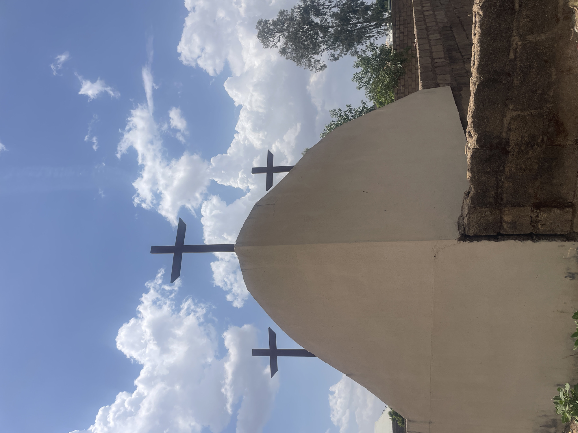 The image shows the Martyrs' Cathedral Church in Jos, Nigeria, under a sky with scattered clouds. Three crosses are mounted on the curved facade of a white building, with sections of a brick building partially visible on the right side. Green foliage is visible near the base of the brick building and possibly on the main structure. Trees are visible in the background. The angle suggests the picture was taken from a low vantage point, looking upwards.