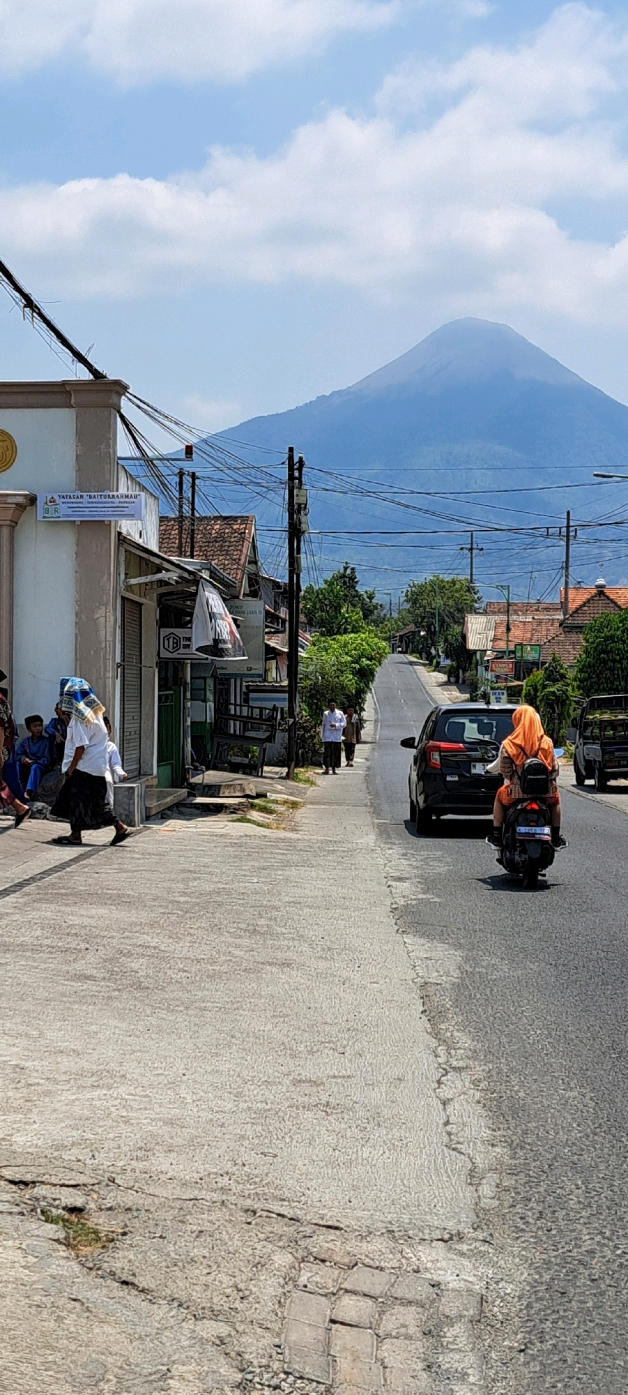 The image captures a street scene in Sumber Gedang, Indonesia, with a prominent volcano in the background. A paved road runs through the middle of the frame, with a concrete sidewalk on the left. Several people are visible on the sidewalk, some walking and others sitting. A woman wearing an orange hijab is riding a motorcycle down the road. Several buildings line the street, including a white building with a sign that reads "YAYASAN DAITURRAHMAH". The sky is partly cloudy. The overall impression is one of a typical day in a semi-urban area with a strong natural backdrop.