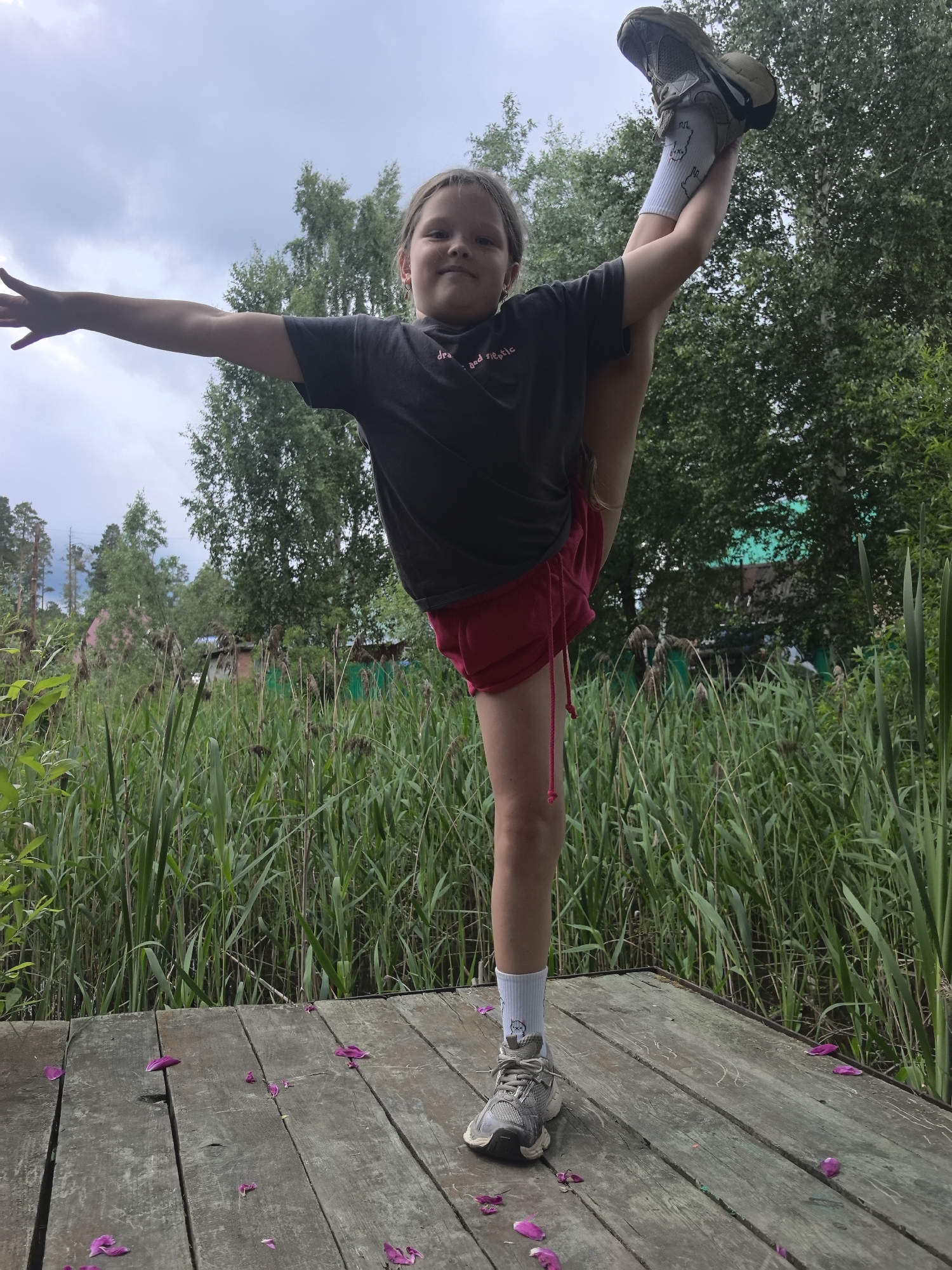 A young girl performs a split with her right leg extended upwards, on a wooden platform surrounded by tall reeds and greenery. She is wearing a dark grey t-shirt and red shorts, white socks with a cartoonish design, and grey athletic shoes. Her left arm is extended horizontally to her left, and her right arm is bent at the elbow, supporting her lifted leg. The girl is looking directly at the camera with a slight smile.
The scene appears to be outdoors, with a cloudy sky visible above the trees in the background. The presence of reeds suggests a natural, possibly marshy or lakeside environment. The wooden platform could be a pier or a boardwalk. Scattered pink flower petals on the wooden planks add a touch of color to the scene. The overall impression is one of athleticism and a connection with nature. There are no clearly visible words on the girl's clothing, except for a small, partially obscured graphic on her t-shirt. The time of day appears to be daytime, given the natural light. The weather seems overcast, indicated by the clouds.