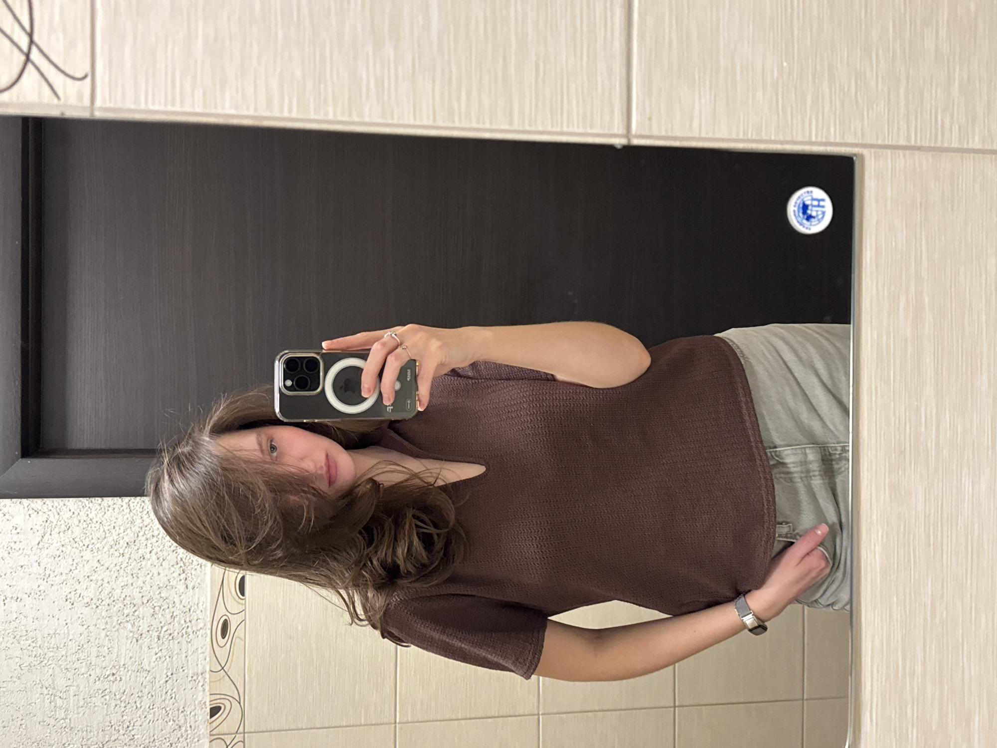 A selfie taken in a bathroom setting. The person in the image is a woman with light skin and brown hair, wearing a brown short-sleeved top and grey pants. She is holding a smartphone in her right hand, presumably to take the picture. The background consists of beige tiles with decorative patterns. There's a dark brown door or panel visible within the mirror's reflection, and a circular sticker with a blue logo is affixed to the mirror. The location is Perm, Russia.