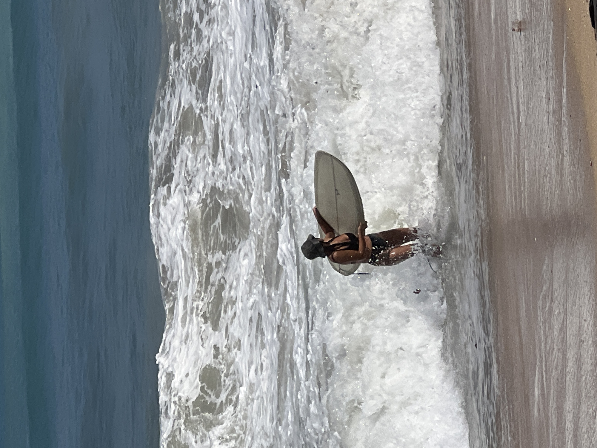 In Kuta, Indonesia, a person wearing a black bikini and a dark bucket hat stands in the ocean holding a surfboard. The person is facing away from the camera, with the surfboard held under their right arm. They are partially submerged in foamy white water created by breaking waves. The ocean water is a deep blue, transitioning to white foam near the shore. The beach appears to be sandy.