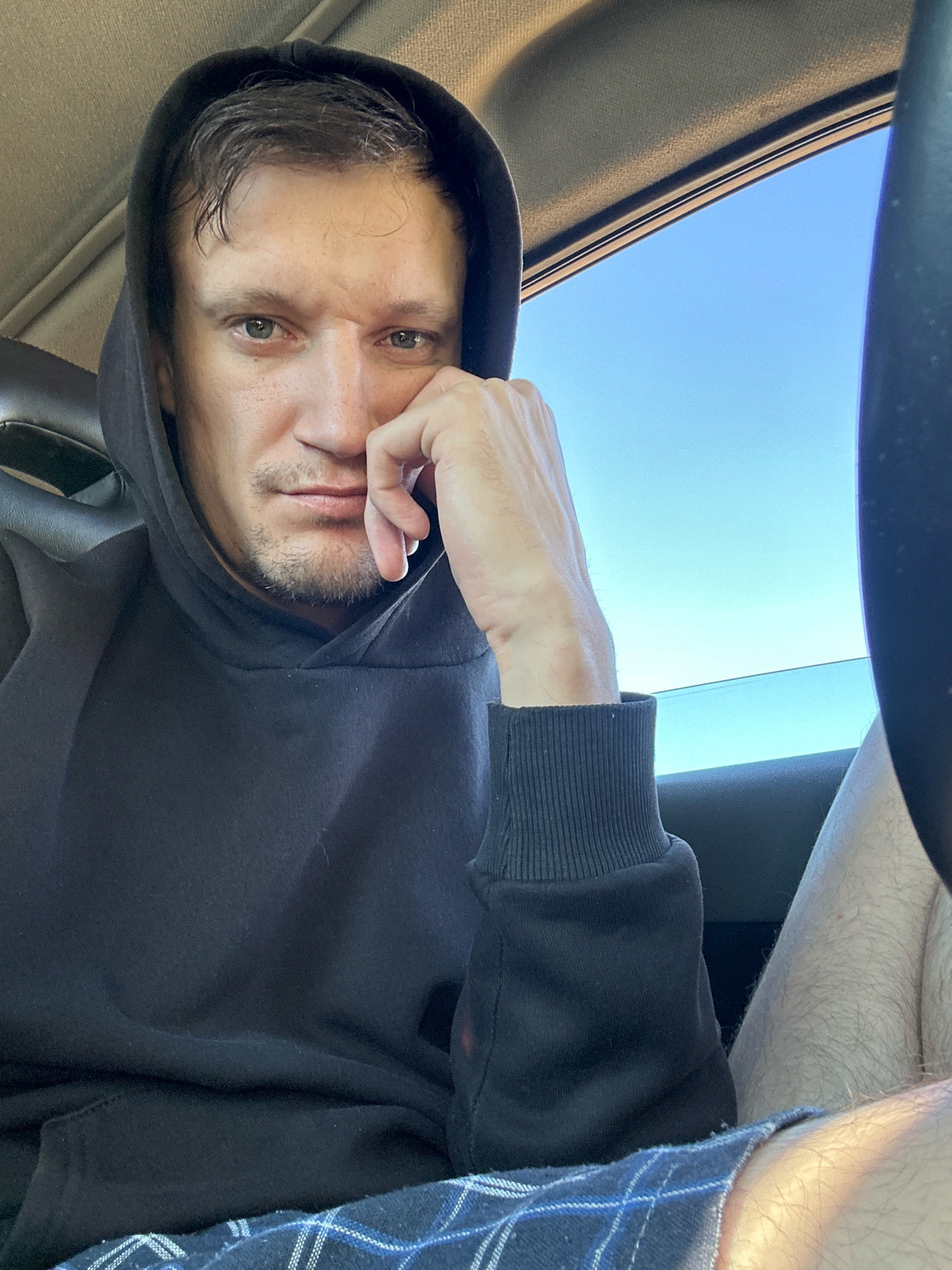 A man wearing a black hoodie and blue checkered shorts is sitting in a car. His right hand is resting on his chin, and he is looking forward with a serious expression. The car's interior is visible, including the car seat and the roof lining. Outside the car window, the blue sky is visible. The image does not provide any information about the specific location in the city of Makiv, Ukraine.