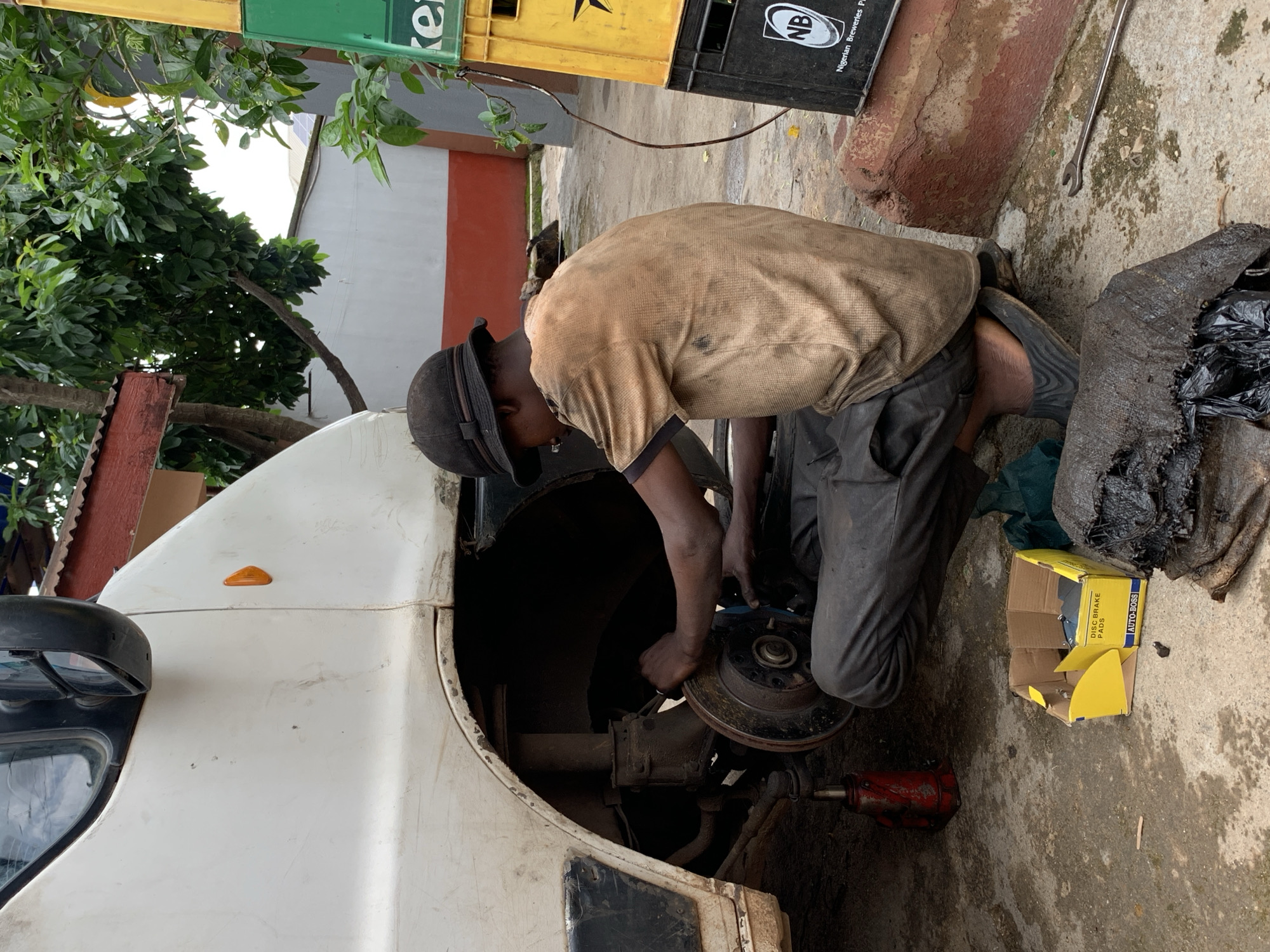 A mechanic is working on the front wheel assembly of a white vehicle. The mechanic is wearing a brown t-shirt, dark pants, and a black cap. He is kneeling on the ground, reaching into the wheel well. A red hydraulic jack is visible beneath the vehicle, and a yellow cardboard box with the text "AUTO-BOSS DISC BRAKE PADS" is on the ground nearby. To the right of the box is a dark, bulky object, possibly a sack of waste or old car parts.
The scene appears to be an outdoor workshop, with a concrete floor that is wet in places, suggesting recent rain or cleaning. In the background, there are some green trees, a red and white striped building, and stacked crates, one yellow and one black with "NB Nigerian Breweries Plc" written on it. A silver wrench lies on the concrete. The lighting suggests it is daytime, though the exact time is unclear. The overall impression is of a busy, functional repair area.