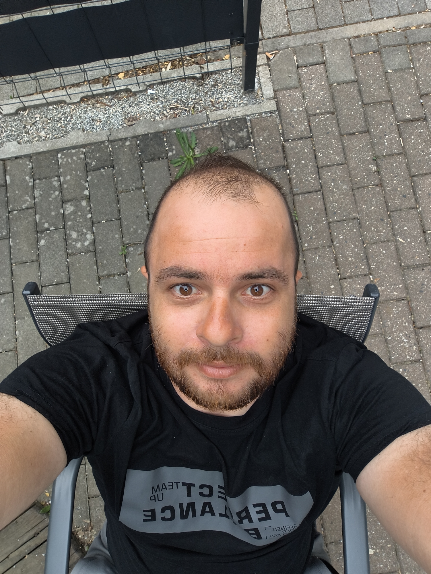 A man with brown hair and a beard is sitting in a chair outdoors. He is wearing a black t-shirt with the words "Team up Performance" printed on it in grey, inverted text. The chair has a grey frame and a patterned backrest. The ground is paved with grey brick, and there is a patch of gravel and a black fence in the background. The location appears to be a patio or garden. The city is Unknown, Unknown.