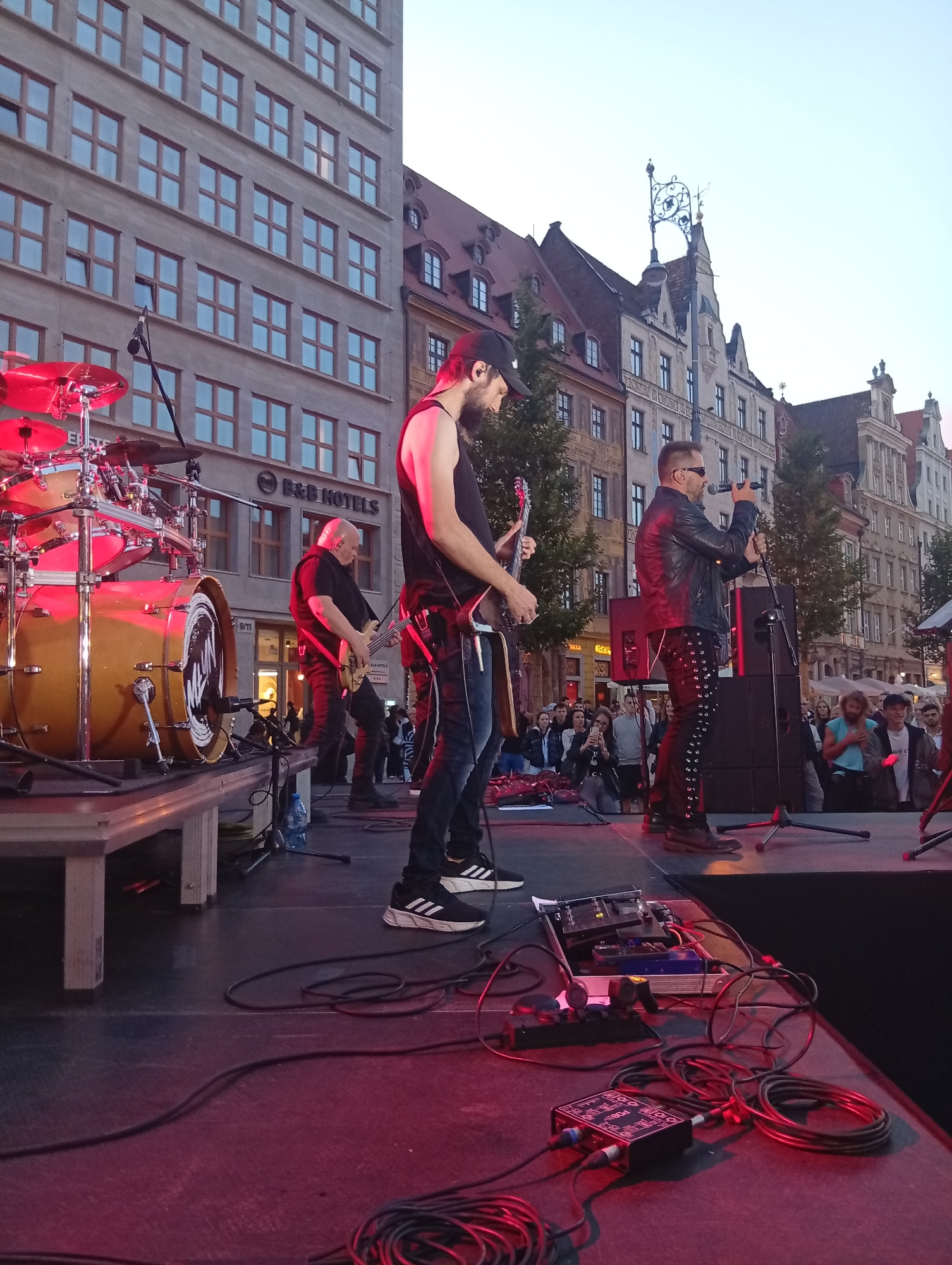 An outdoor concert is taking place on a stage in Wrocław, Poland. The stage is set up in front of historic buildings, with a tall, modern building to the left and older, ornate buildings to the right. A band is performing, with a guitarist in the foreground on the left, a bassist in the middle, and a singer on the right. The singer is wearing a black leather jacket and pants adorned with silver eyelets, and sunglasses. The band members are playing their instruments and singing into microphones. The stage is equipped with musical instruments, amplifiers, and audio equipment. A crowd of people is gathered in front of the stage, watching the performance. The scene is illuminated by stage lights, casting a red glow on the stage and the performers. The sky is visible in the background, with a light overcast.