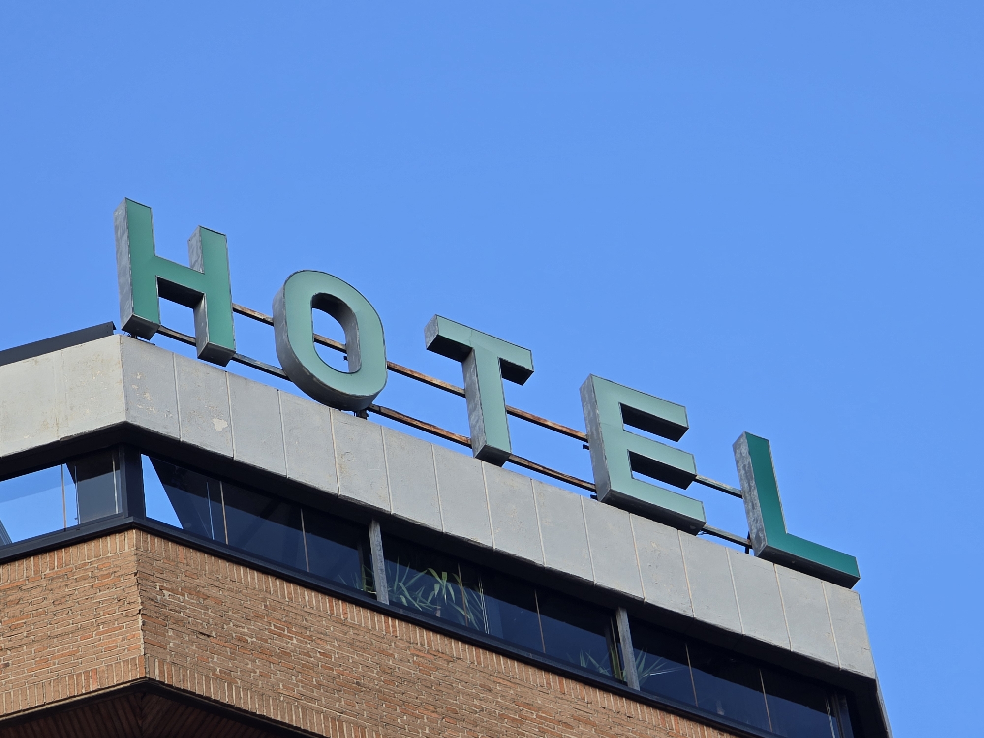 The image shows a close-up, low-angle shot of a building with the word "HOTEL" spelled out in large, green, three-dimensional block letters on its rooftop, against a clear blue sky. The building has a brick facade on the lower section, with a row of windows above. The upper section of the building, supporting the sign, is made of a gray material. The letters are mounted on metal supports. There are no people visible in the image. The scene is an exterior view of a hotel sign in an urban setting, specifically located in the city of Jaén, Spain.