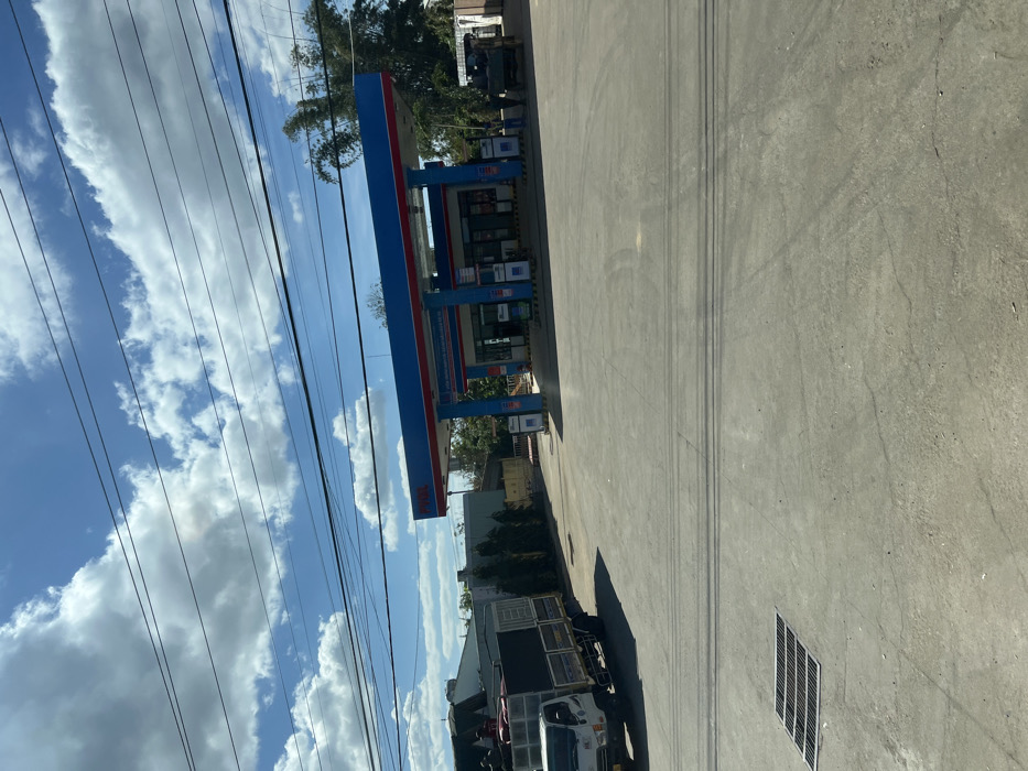 A gas station is visible under a bright blue sky with scattered clouds, intersected by power lines. The station has a prominent blue and red canopy, with multiple gas pumps underneath. A truck sits near the edge of the frame, and a grated drain is on the paved ground. A building and some trees are behind the gas station, and some distant trees are at the top of the frame.