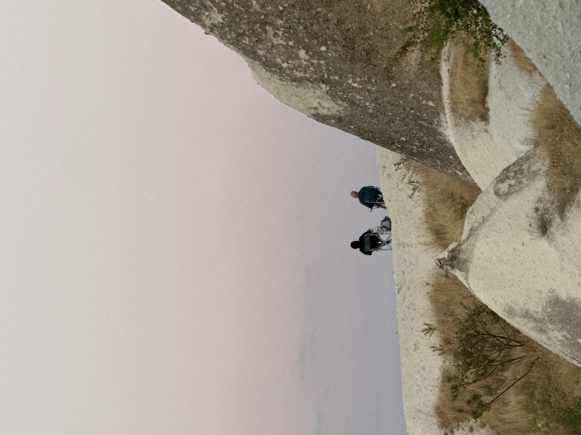 In Göreme, Turkey, two individuals are seated in camping chairs on a sandy ridge, facing away from the camera, with a soft pink and pale purple sky behind them. The person on the left wears a dark hooded jacket and is positioned slightly in front of a backpack. The person on the right, with short hair, wears a dark jacket and is seated slightly behind and to the right of the first person. The landscape is characterized by undulating, pale, sandy mounds and rock formations, with dry grasses and sparse vegetation dotting the scene. A large, textured rock formation dominates the right side of the image, extending upwards.
