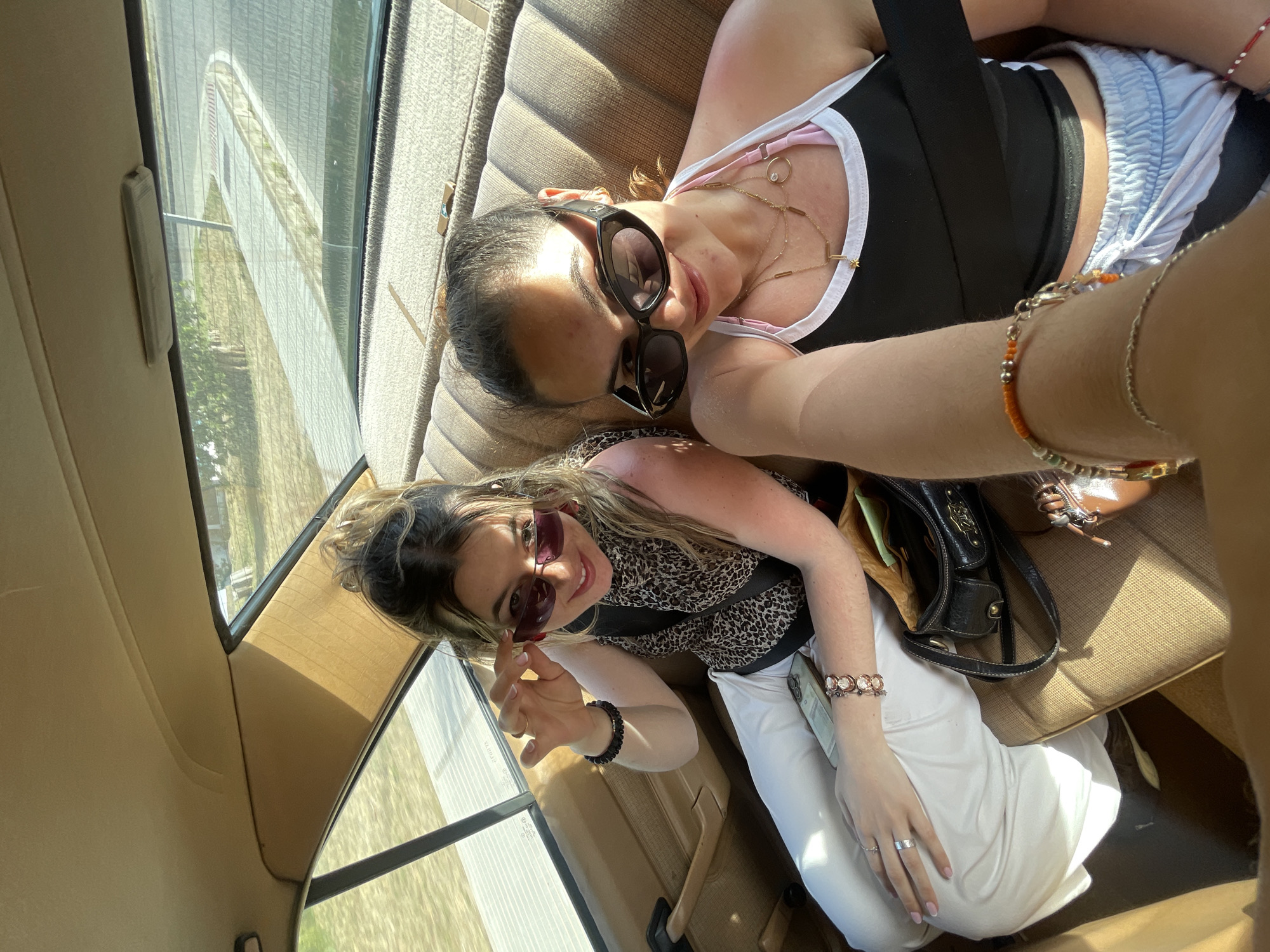 Two young women are inside a vehicle, possibly a car, taking a selfie. The vehicle appears to be moving, based on the blurred scenery visible through the windows. One woman is wearing a black tank top with a white trim and blue shorts, accessorized with multiple necklaces and bracelets. She is wearing large, dark sunglasses. The other woman is wearing a patterned blouse, white pants, and sunglasses. She has several bracelets on her wrist. The interior of the vehicle is beige. There are purses present. The exterior view suggests a road or highway setting. Based on the clues, it is not possible to pinpoint the exact location in El Puerto de Santa María.