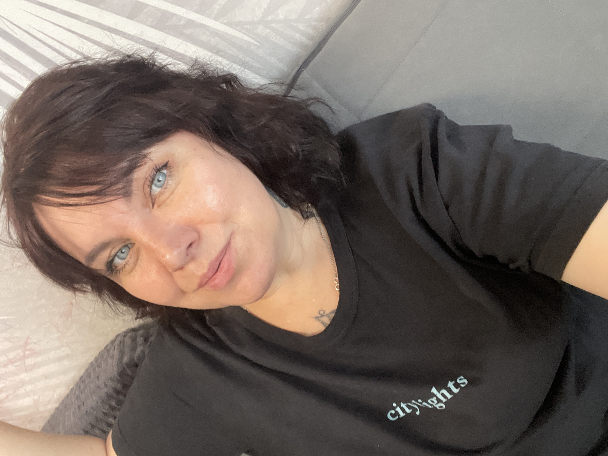 A middle-aged woman with short, dark, wavy hair and striking blue eyes is captured in a close-up selfie. She is wearing a black t-shirt with the text "city lights" in light blue lettering. Her left arm is bent and resting behind her head, and she is looking directly at the camera with a slight smile. The background consists of a textured wall with a light-colored, geometric pattern, and a grey sofa. There is a subtle hint of what appears to be a tattoo on her collarbone. The lighting is soft and natural, suggesting an indoor setting. There is no indication of the specific location in Zhukovo, Russia.