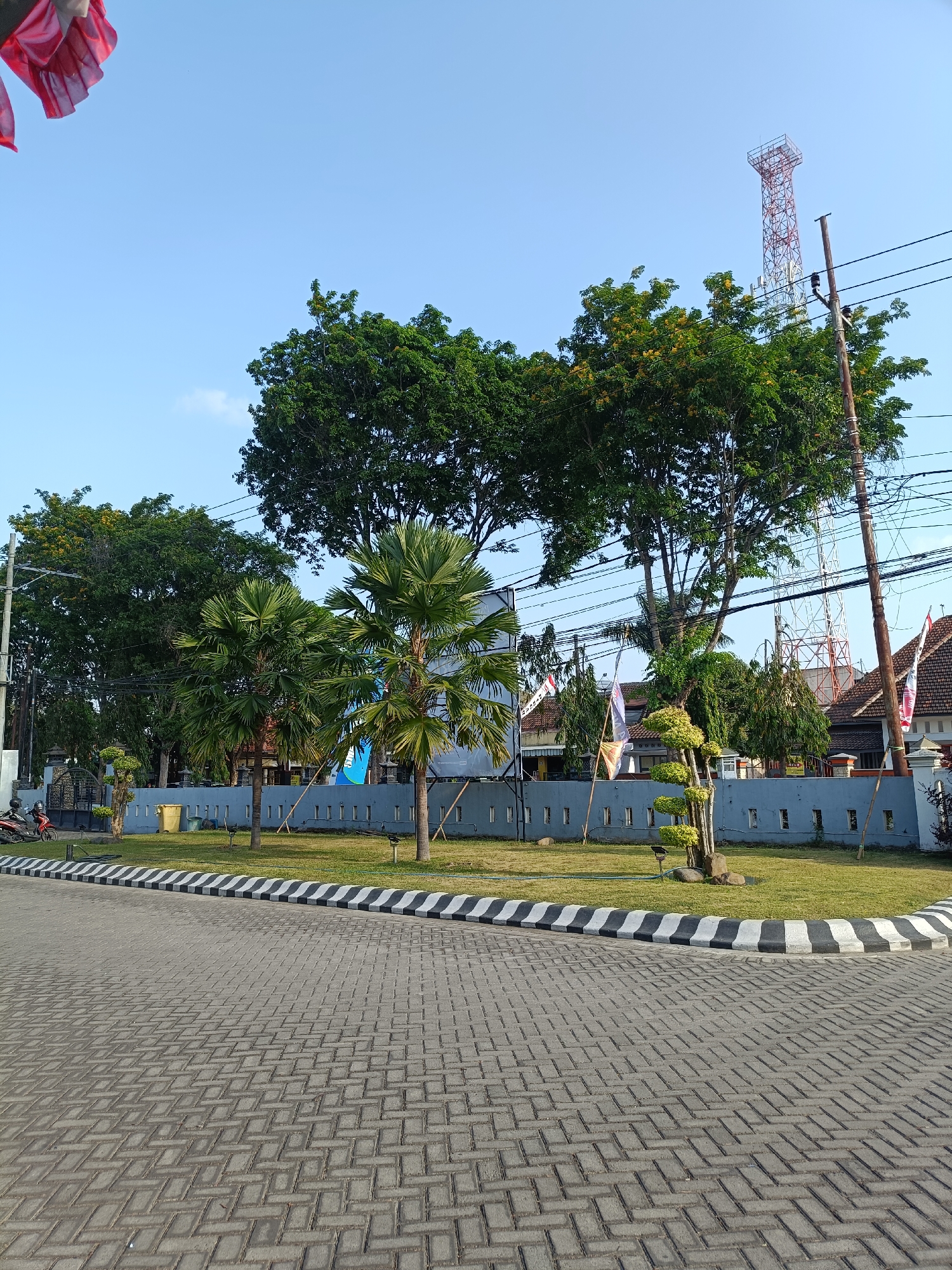 The image depicts a paved area with brick-like patterns in the foreground, leading to a landscaped section with green grass. A curved border with alternating black and white stripes separates the pavement from the grass. Beyond the grass, a grey wall with rectangular indentations runs horizontally. Two palm trees and a sculpted tree stand in the grassy area. In the background, a collection of buildings with tiled roofs can be seen, along with a red and white lattice tower and utility poles with wires. Several flags are visible, including one red and white Indonesian flag. A motorcycle is parked on the left side of the image. The sky is clear and blue. The scene appears to be an outdoor public space, possibly in a residential or commercial area, given the presence of buildings and utility infrastructure. The presence of the Indonesian flag suggests the location is within Indonesia. The combination of a paved area, landscaping, buildings, and communication towers is common in urban environments.