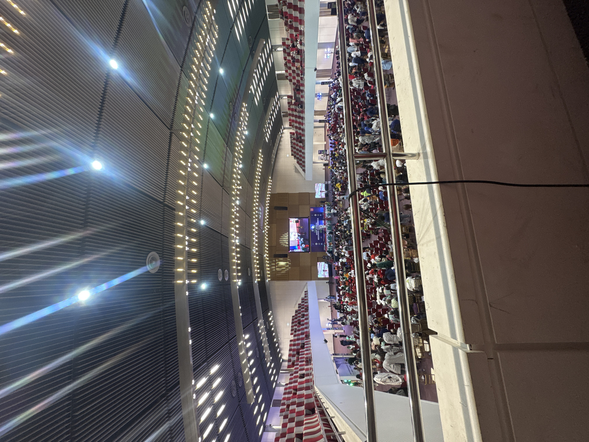 The image, originally rotated 90 degrees clockwise, provides an elevated view inside a large, brightly lit indoor conference hall or auditorium in Abuja, Nigeria. The hall is filled with a substantial audience, with numerous individuals seated in orderly rows of red and some lighter-colored chairs on the main floor.
The ceiling features a distinctive design with horizontal slats or ribs and multiple recessed lights that cast bright beams and lens flares across the surface. In the foreground, a metal railing marks the edge of an upper viewing level, with a black cable visible draped over it.
At the far end of the hall, several large digital screens are prominent. One screen clearly displays the text "WOMEN AFRICA" and "CONFERENCE," identifying the event taking place. Another screen shows what appears to be a logo or additional text, including fragments like "WORLD AFRICA." Below the screens, there are sections with more seating and what might be a stage or presentation area.
A decorative architectural element featuring a red and white checkered pattern is visible along a lower wall or partition running parallel to the main seating section. The scene depicts a large-scale gathering, likely a conference or convention, with attendees observing the presentations on the screens. The event is well-illuminated by artificial lighting within the spacious venue.