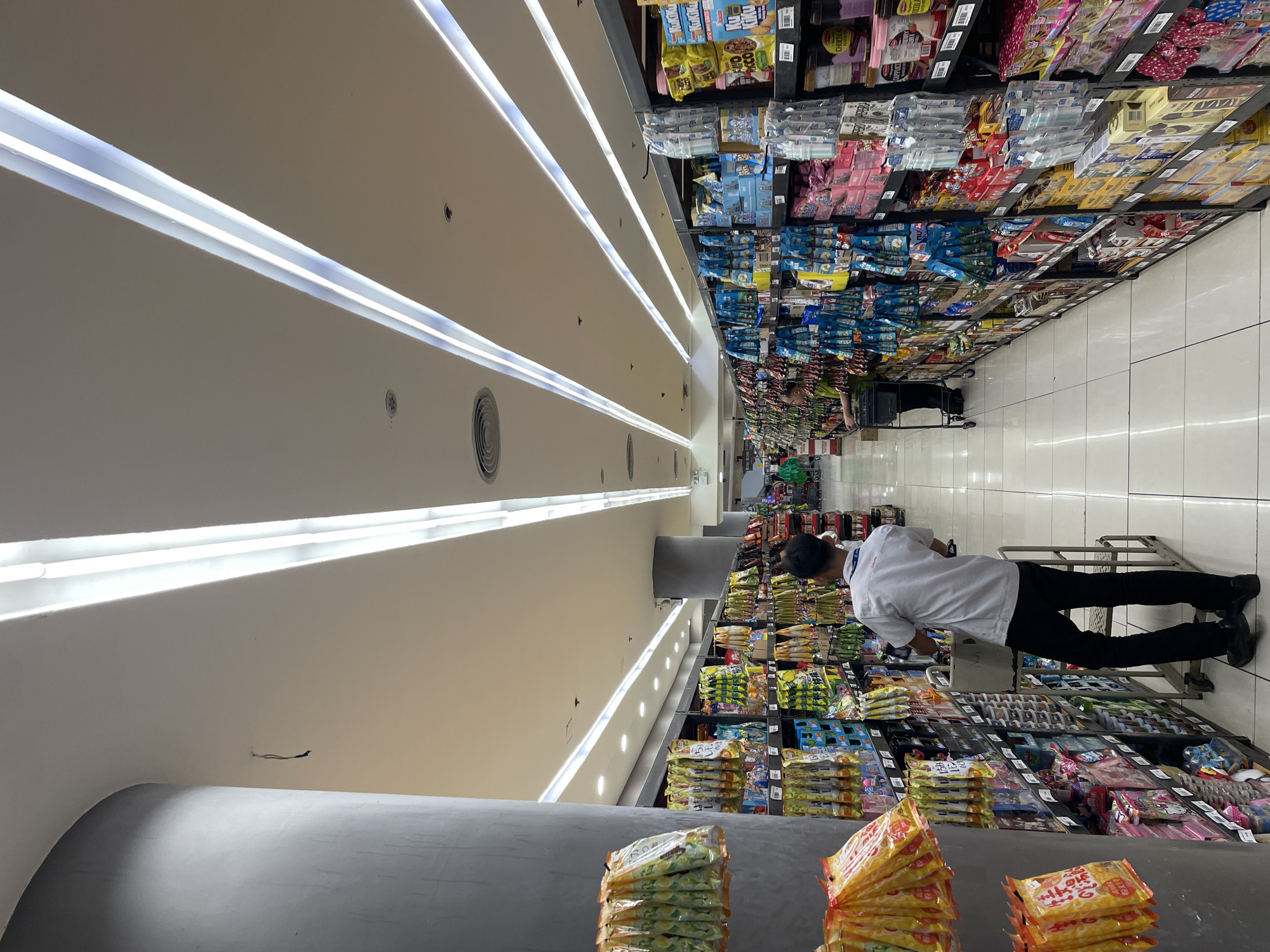 The image shows the interior of a supermarket aisle in Davao City, Philippines. The aisle is well-lit with long, fluorescent ceiling lights. Shelves on both sides are stocked with various packaged goods, primarily snacks and food items. A person wearing a white polo shirt and black pants, likely a store employee, is seen from behind, pushing a shopping cart. Another person is partially visible further down the aisle, also with a shopping cart. The flooring appears to be tiled. The overall impression is a typical retail environment stocked for customers.