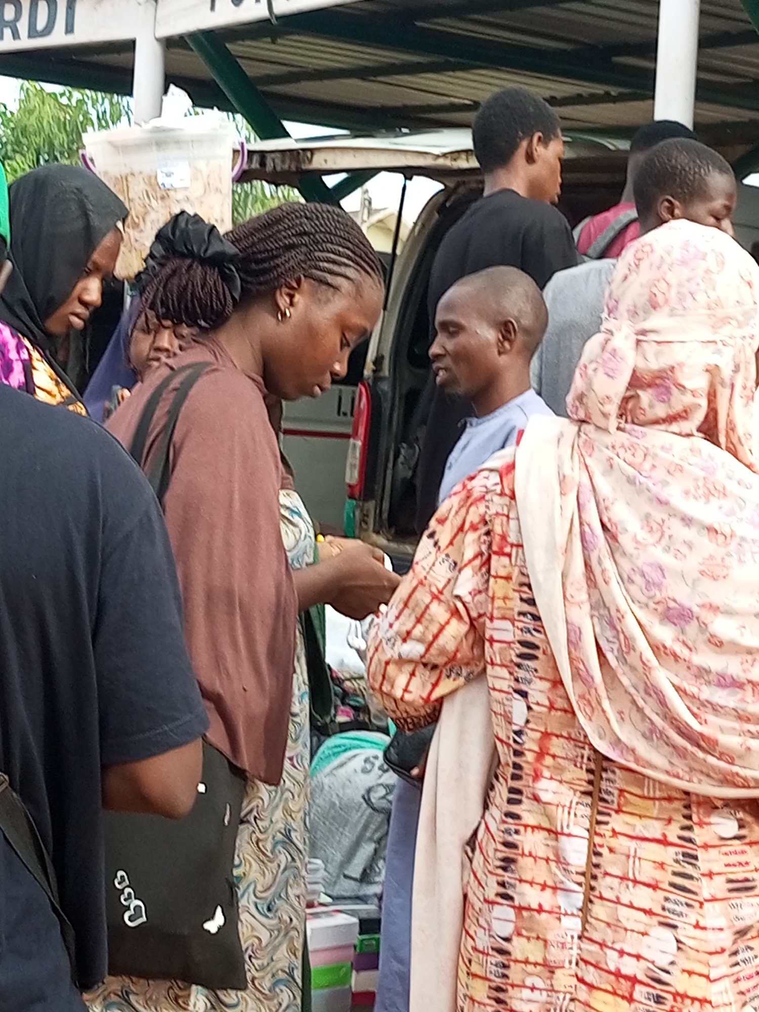 The image depicts a street scene, likely a marketplace, in Gombe, Nigeria. Several individuals are visible, predominantly women, dressed in traditional African attire. One woman in the foreground, wearing a brown headscarf and a dark brown top, is focused on something she is holding in her hands. To her left, another woman in a black hijab looks down. To the right of the first woman, another individual, possibly a man, is partially visible, wearing a light blue shirt. In the background, other people are seen, including a person wearing a pink garment. The environment appears to be outdoors, with a covered structure overhead, possibly a market stall or shelter. The overall impression is that of a busy, everyday scene in a Nigerian urban setting.