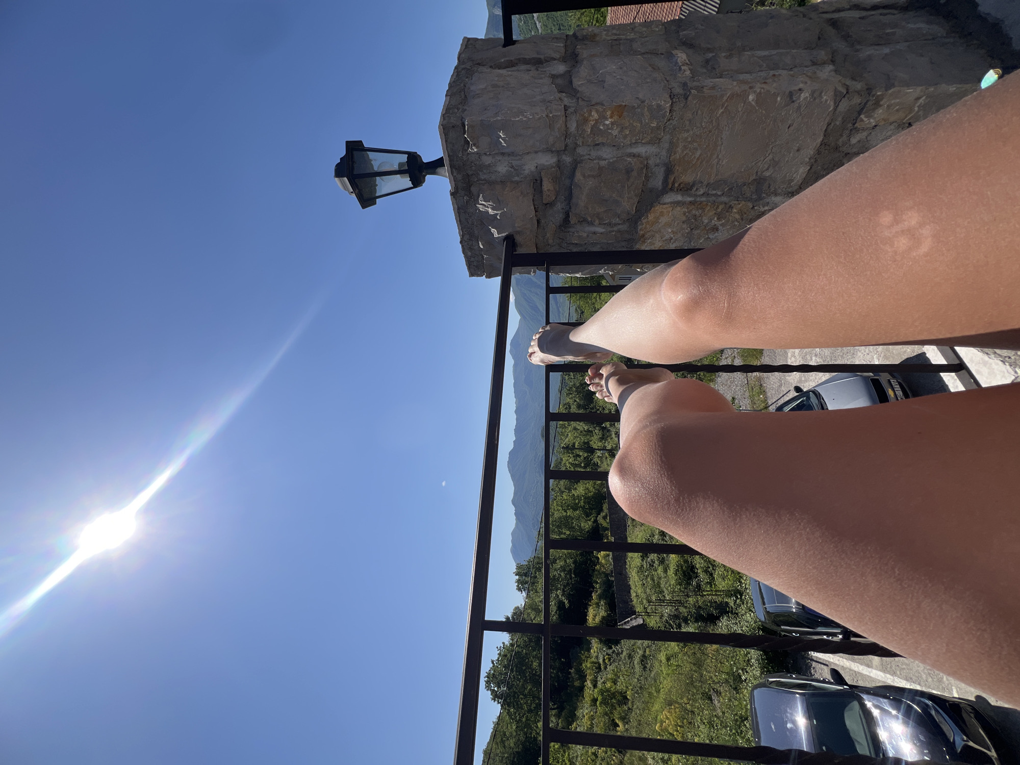 The image shows a view from a balcony or elevated platform, likely in Sotonići, Montenegro, based on the terrain and architecture. A person's legs are prominently featured, resting on the balcony railing. The railing is dark metal. Beyond the railing, there is a view of trees, distant mountains, and a clear blue sky. The sun is visible in the sky, creating a lens flare effect. There is a stone pillar to the right with a lamppost attached. Several cars are parked underneath the view.