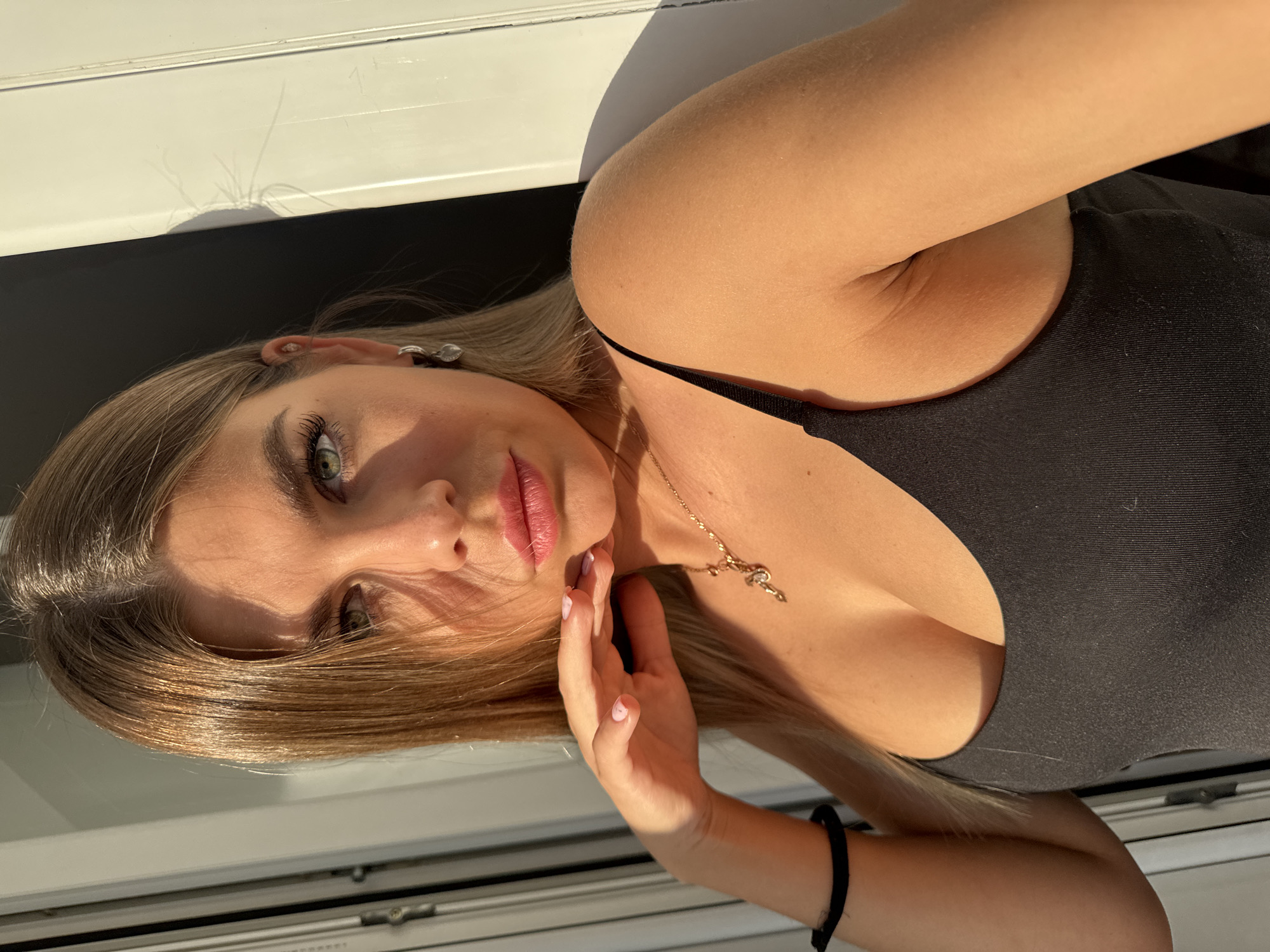 The image shows a close-up of a young woman with blonde hair and green eyes, posing indoors. She is wearing a black tank top and a gold necklace. Her hand is resting on her chin, and she is looking directly at the camera. The lighting suggests it is daytime, with sunlight casting shadows on the wall behind her. The background includes part of a window frame and a door. There is no indication that this image is located in Sarapul, Russia.