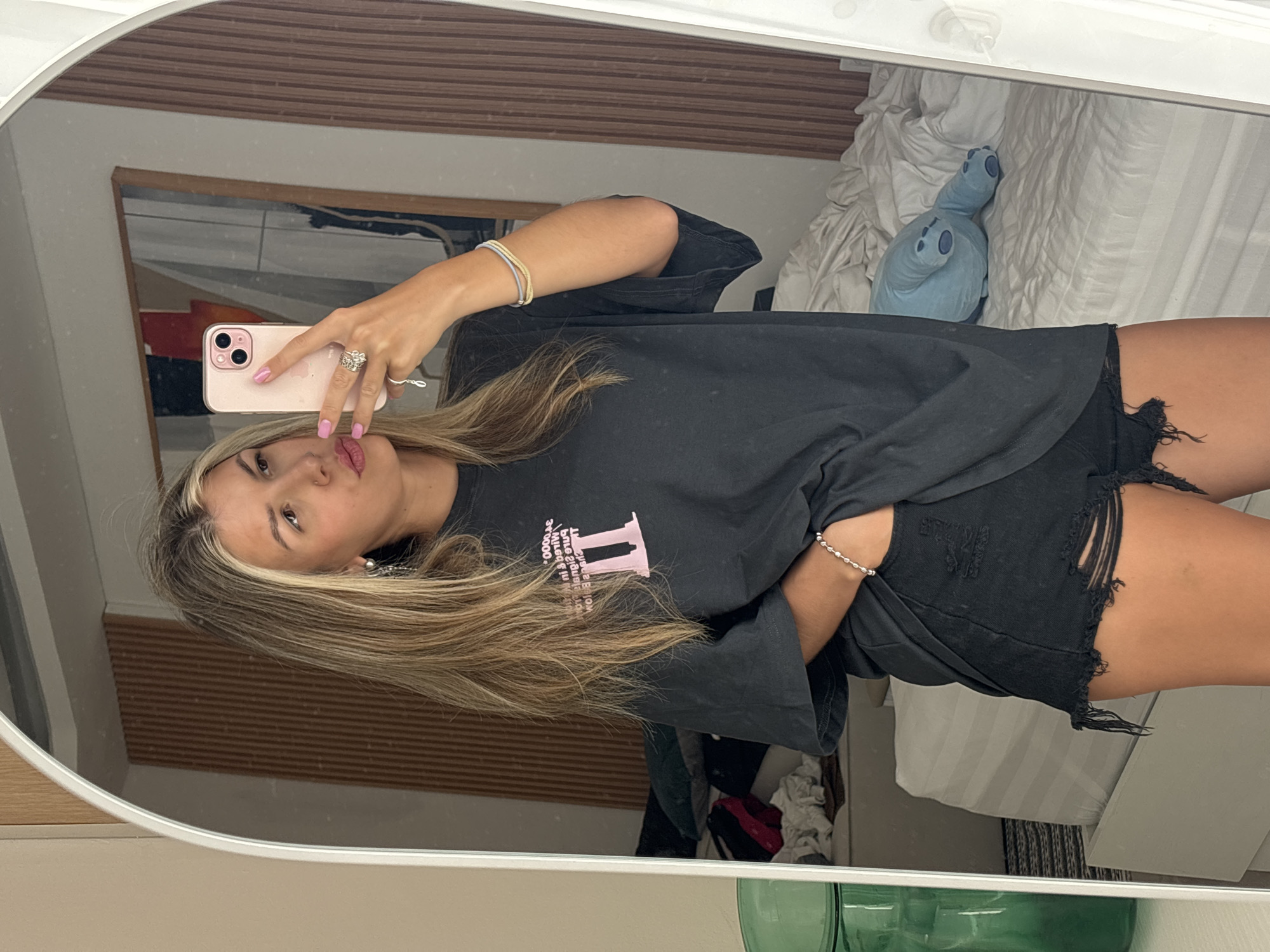 A woman is taking a mirror selfie in what appears to be a hotel room. She is wearing a black t-shirt with pink text and the Roman numeral "II" and black distressed denim shorts. She has blonde hair and is wearing jewelry including earrings, a ring, and bracelets. In the reflection of the mirror, a messy bed with a blue stuffed animal is visible, along with a wooden-paneled wall and artwork. A green glass vase is also visible to the left of the mirror.