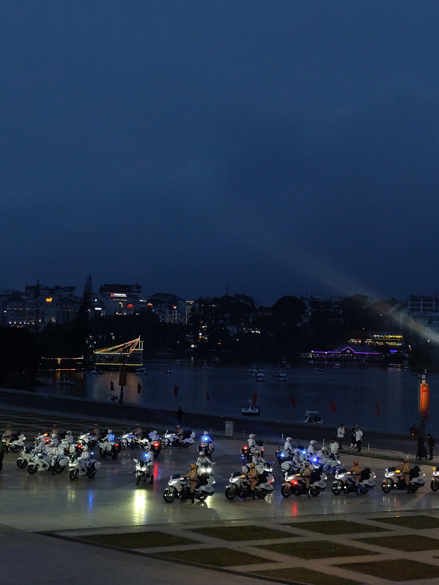 This media file captures a night scene in Da Lat, Vietnam, likely during a ceremonial event or parade. The foreground features a formation of uniformed individuals on motorcycles, many of whom appear to be law enforcement officers, as indicated by their attire and the style of the motorcycles. The motorcycles are predominantly white, with some displaying flashing lights in red and blue, suggesting a display of authority or an organized procession. The riders are clad in helmets and uniforms, some in white and others in brown or khaki.
The middle ground shows a body of water, possibly a lake or river, reflecting the lights from the surrounding area. A decorated boat, adorned with colorful lights, is visible on the water, adding to the festive or ceremonial atmosphere. Several smaller boats, resembling swan pedal boats, are scattered across the water.
In the background, the Da Lat cityscape is illuminated, with buildings showcasing a variety of lights, including neon signs. The overall ambiance is one of twilight or early night, with the sky a deep, dark blue. The lighting suggests artificial illumination is prominent, with spotlights or beams of light visible in the upper right portion of the image, possibly from a performance or display.
The event appears to be a public gathering or demonstration, given the organized presence of the motorcyclists and the illuminated cityscape. The presence of a boat with decorative lights and the reflective water create a picturesque and potentially celebratory mood. No specific emotions are evident from the individuals due to the distance and lighting, but the organized nature of the scene implies order and perhaps a sense of occasion.
Visible text is limited but one of the motorcycles in the foreground has what appears to be "C7GT" written on its side. Another visible sign is a vertical banner on the far right of the image, red with some white text that is too small to decipher clearly. The overall scene depicts a lively urban setting at night, with a focus on an organized procession of motorcycles and the illuminated waterfront.