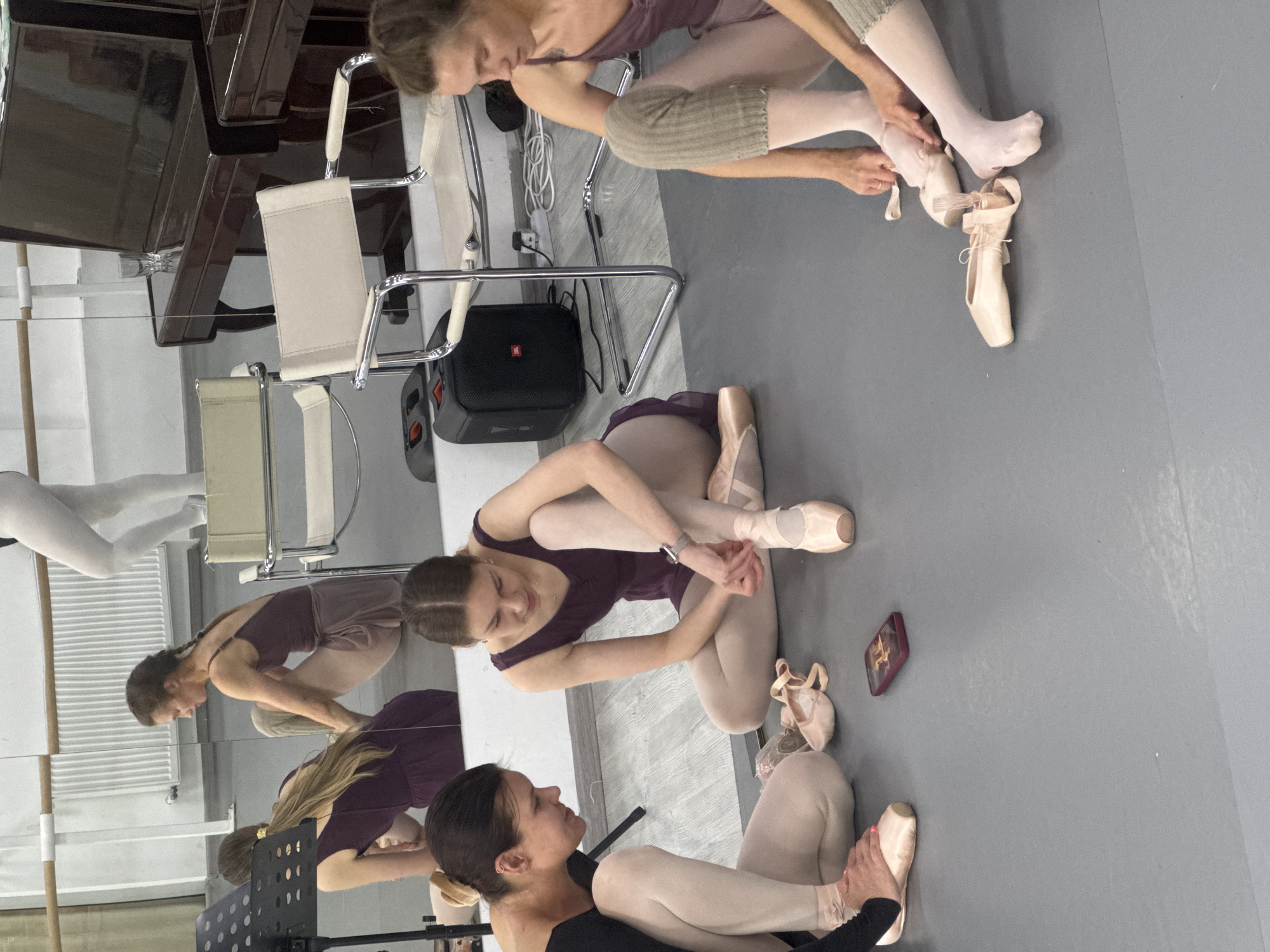 Three ballerinas are seated on the floor of a dance studio, preparing their pointe shoes. The room has a gray floor and mirrored walls. A black speaker and chairs are present. The location is likely in Tbilisi, Georgia.