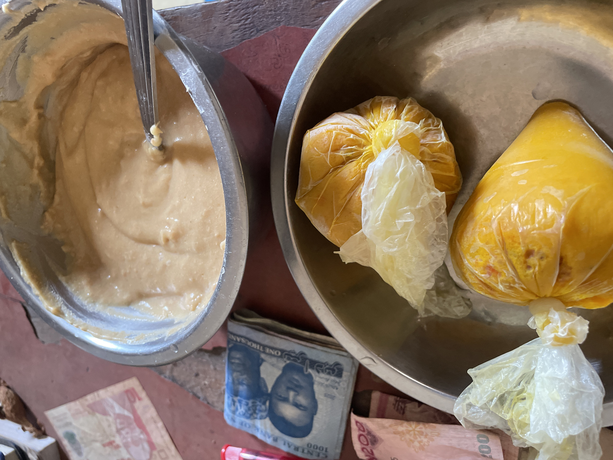 This image, captured in Onitsha, Nigeria, displays an arrangement of food items and currency on a reddish-brown floor or low-lying surface.
In the upper-center, a stainless steel bowl contains a light brown, viscous, semi-liquid substance, consistent with a food paste or batter. A metal spoon or utensil is partially submerged within this substance, positioned towards the right side of the bowl.
To the lower-center, another stainless steel bowl holds several items individually wrapped in clear plastic. These bundles contain a vibrant yellow or orange-colored substance, likely food, with at least two distinct bundles visible.
To the left of the lower bowl, a folded stack of Nigerian Naira notes is present. The top note is clearly identifiable as a 1000 Naira bill, displaying portraits and "ONE THOUSAND" text. A pinkish 200 Naira note is partially visible beneath it. A small red cylindrical object, possibly a pen cap, is situated near the currency. No individuals are visible in the frame, and no direct interactions are depicted. The scene suggests food preparation, sale, or storage in a domestic or small commercial setting.
