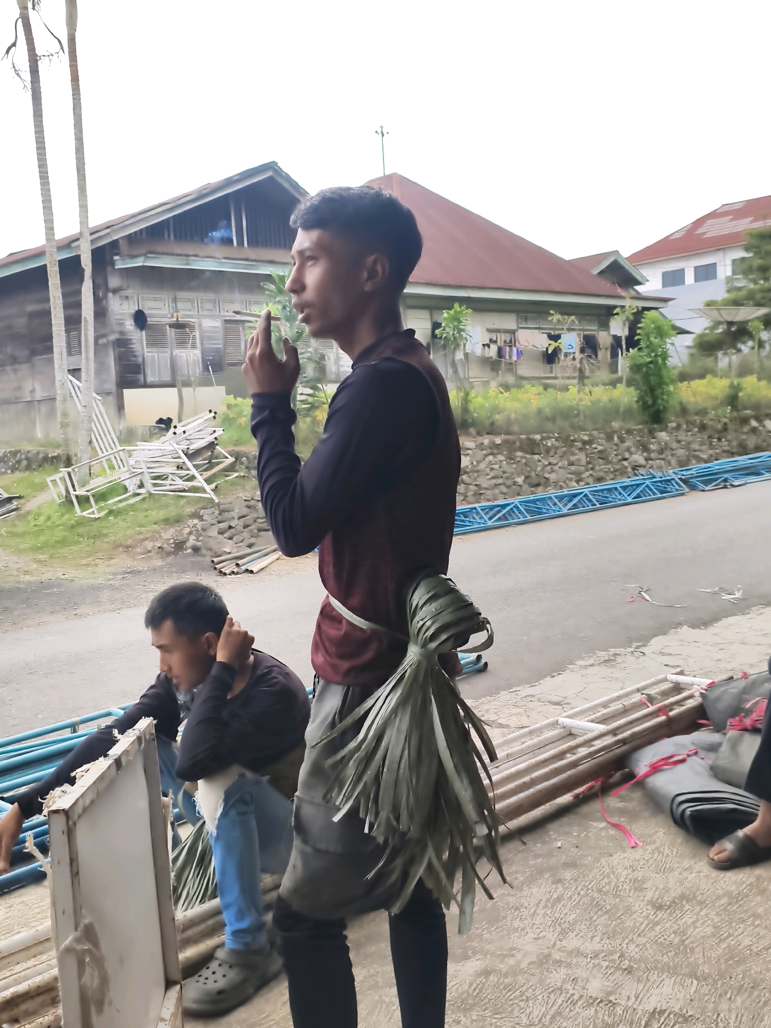 In Lindung Jaya, Indonesia, two men are visible in the foreground. One man stands facing right, smoking a cigarette and wearing a dark long-sleeved shirt with a maroon overlay and a light green sash. A bundle of green leafy material is tied around his waist. The second man kneels facing left, wearing a dark long-sleeved shirt and blue jeans, with one hand touching his head. He is seated on a white metal structure next to a white panel.
In the background, a narrow paved road runs horizontally. On the left side of the road, a weathered wooden house with a red roof and open shutters stands behind a grassy area with scattered white metal structures. On the right side of the road, a row of blue metal truss sections lies on the ground, and beyond that, a modern building with a red roof is visible. The sky is overcast. Scattered around the scene are various metal structures, bundles of material, and coiled grey fabric.