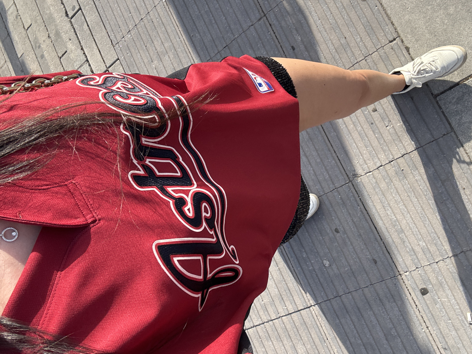 The image shows a person wearing a red baseball jersey with "Astros" written across the chest in black and white lettering. They are also wearing black sequined shorts and white sneakers. The person is standing on a sidewalk made of grey rectangular tiles. The image appears to be taken outdoors on a sunny day. The location is not specified in the image, and therefore cannot be determined to be Madrid, Spain.