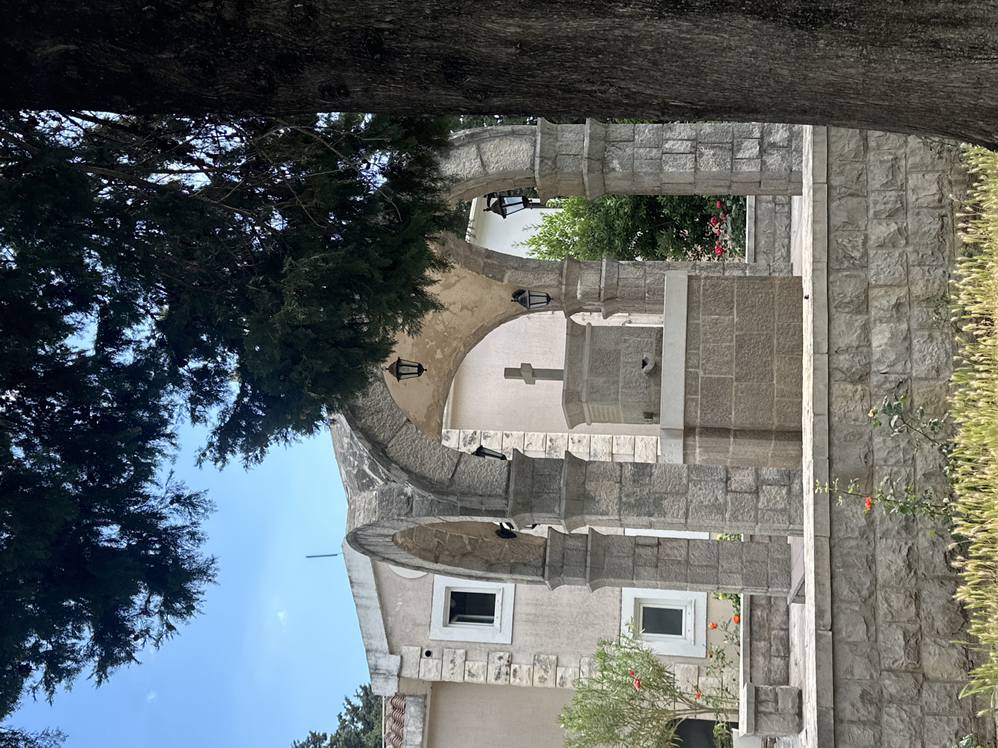 The image depicts an outdoor scene in Boreti, Montenegro. It showcases a building with an architectural design that includes arches, stone construction, and small rectangular windows. A large tree and its foliage frame the scene on the left and in the upper portion of the image, adding a natural element. A stone wall with some vegetation growing on it is visible on the right. A cross is visible on the wall of the building. There are also old-fashioned lanterns installed under the arches of the building. The lighting suggests that it is daytime.