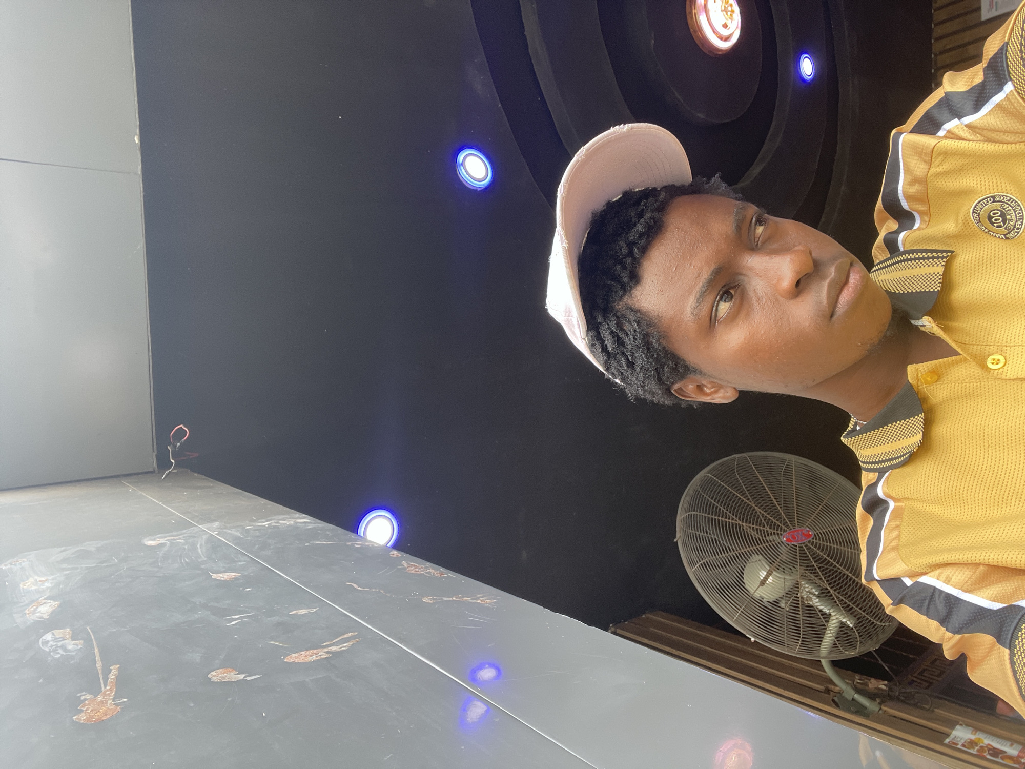 In Nkpor, Nigeria, a young Black male, wearing a light pink baseball cap and a bright yellow polo shirt with black and white accents, is captured in a close-up shot from the chest up. He is looking to his right, his expression contemplative. The background is a dark ceiling with several small, circular blue lights and one larger, warmer light fixture visible. To the left, a large industrial fan is mounted on a wall, with a portion of a textured, dark grey wall featuring reddish-brown abstract markings above it. The lighting suggests an indoor setting, possibly a commercial or public space, as indicated by the prominent fan and overhead lighting. The overall ambiance is casual and observational.