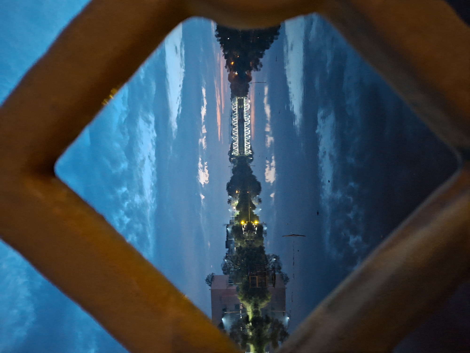 The image shows a view through a diamond-shaped metal structure of a river in Lhokseumawe, Indonesia, at dusk. The sky is a deep blue with streaks of orange and pink from the setting sun, and clouds are visible. The river is calm and reflects the sky and the lights from the buildings and a bridge on the opposite bank. On the left side of the river, there is a large building and some trees. In the center of the scene, a long, illuminated bridge with a triangular truss structure crosses the river. The reflection of the bridge and the surrounding lights is clearly visible on the water's surface. Trees line the banks of the river, and some smaller structures and lights are scattered along the shoreline. The foreground is dominated by the blurry, golden-colored metal bars of the diamond-shaped structure through which the scene is viewed.
