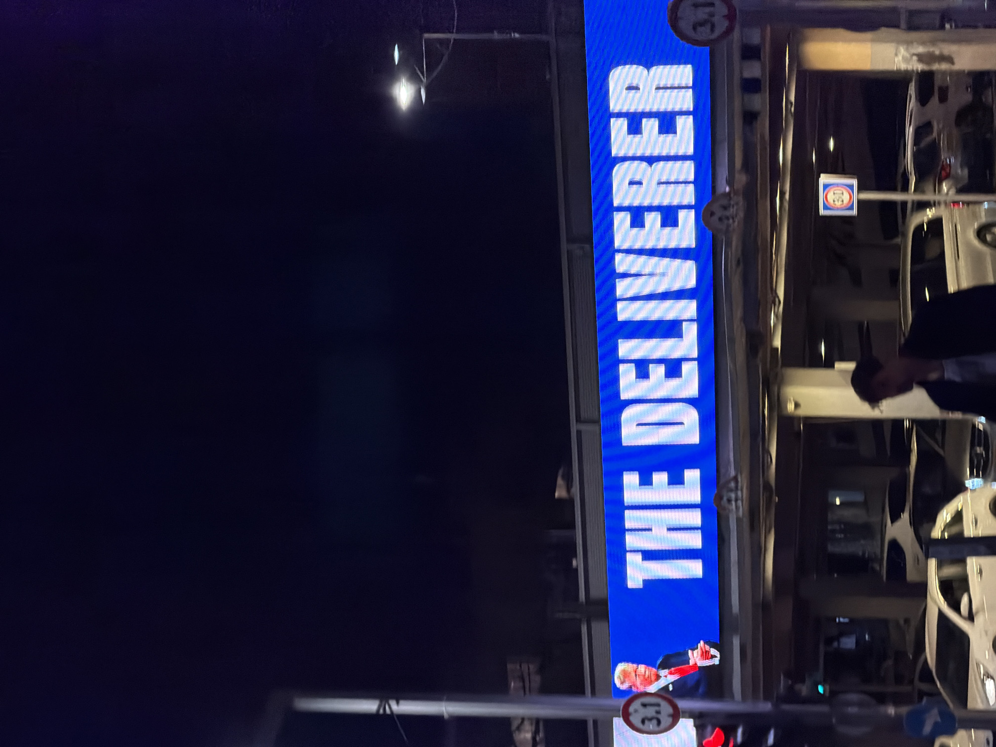 The media file displays a night scene, primarily featuring a large, vertical, illuminated LED billboard. The billboard prominently features the text "THE DELIVERER" in bold, white, capital letters, oriented vertically and reading upwards, against a bright blue background. At the bottom of the billboard, a stylized image of Donald Trump is visible, depicted in a suit and red tie, with his right arm raised in a gesturing pose.
To the left of the illuminated billboard, the dark night sky stretches upwards, with a street light emitting a distinct, star-like glow. Several metal poles and street infrastructure elements are visible throughout the frame. Attached to these structures are two circular red and white signs, each displaying "3.1," indicating a height restriction of 3.1 meters. One sign is located near the lower left portion of the billboard, and another is positioned higher up on the right side of the frame. A smaller square sign with a blue circular symbol is also present further to the right.
The right side of the image reveals reflections of multiple white vehicles, possibly SUVs or vans, suggesting a parking area or street parallel to the billboard, seen through a glass facade or window. The head and shoulders of a person, looking downwards, are visible on the far right, and another partial figure is present at the bottom right. The setting appears to be an urban street or thoroughfare in Tel-Aviv, Israel, captured after dark, with artificial lighting illuminating the scene. No specific activity or emotion is clearly discernible, offering an observational snapshot of the urban landscape.