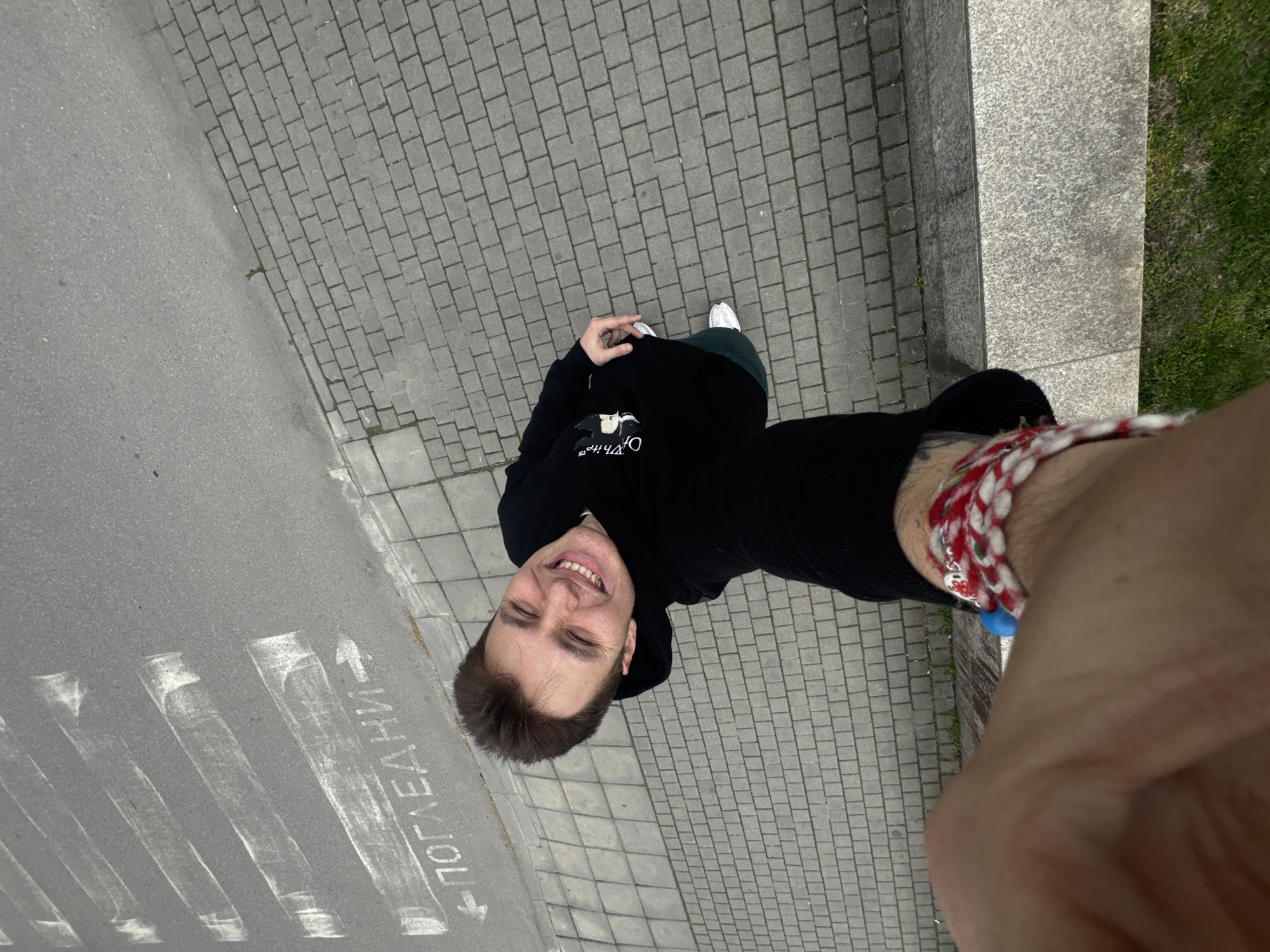 A man is standing on a cobblestone sidewalk next to a street in Plovdiv, Bulgaria, taking a selfie. He is wearing a black hoodie and green pants. His left hand is holding his phone to take the photo, and a red-and-white Martenitsa bracelet is on his left wrist. The street has a crosswalk with the word "ПОГЛЕДАНИ" painted on it. There is a concrete curb and some grass next to the sidewalk.
