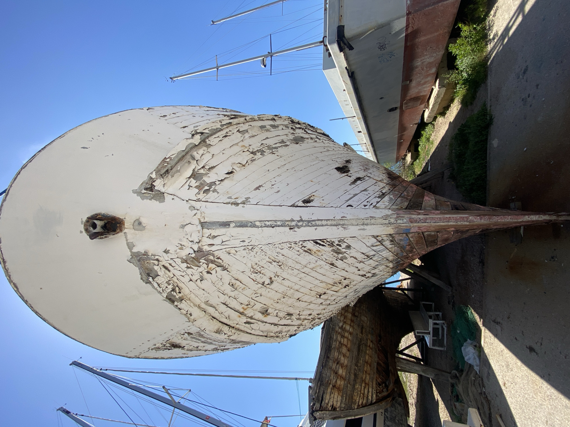 The image depicts the hull of an old, wooden boat in a shipyard in Bar, Montenegro. The boat is white, but much of the paint is peeling off. The wooden planks underneath are visible. It rests on wooden supports above the ground. Other boats are in the background, and the masts of sailboats are seen in the clear blue sky. There are chairs and tarps stowed underneath the wooden hull.
