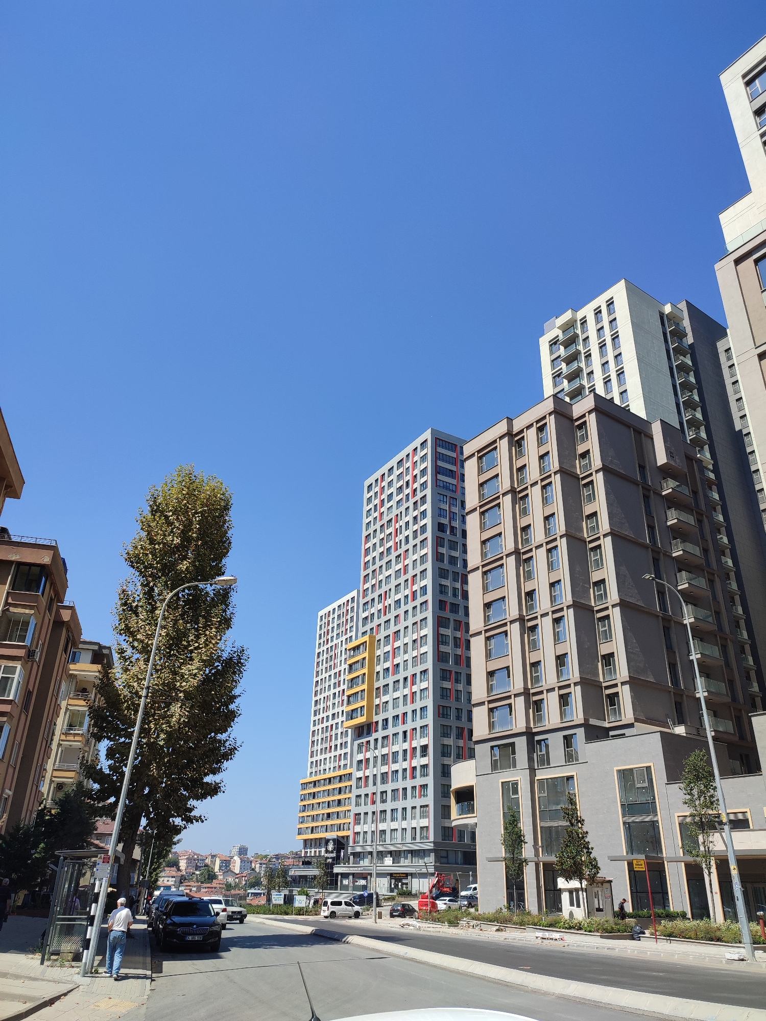 A street scene in Kadıköy, Turkey, under a clear blue sky. Tall residential buildings dominate the right side of the image. The closest building is a modern, mid-rise structure with a grey facade, large windows, and balconies. To its left is a taller, slender building with a white and grey exterior, featuring repeating patterns of windows and balconies. Further left, another tall building stands out with a facade of alternating red and white colored window frames. In the foreground, a street runs horizontally, with cars parked along the right side. A bus stop is visible on the left, with a man standing nearby. A large, leafy tree with a streetlamp in front of it stands prominently in the mid-ground, partially obscuring the buildings. The background reveals a cityscape with more buildings and a hint of rolling hills under the bright sun.