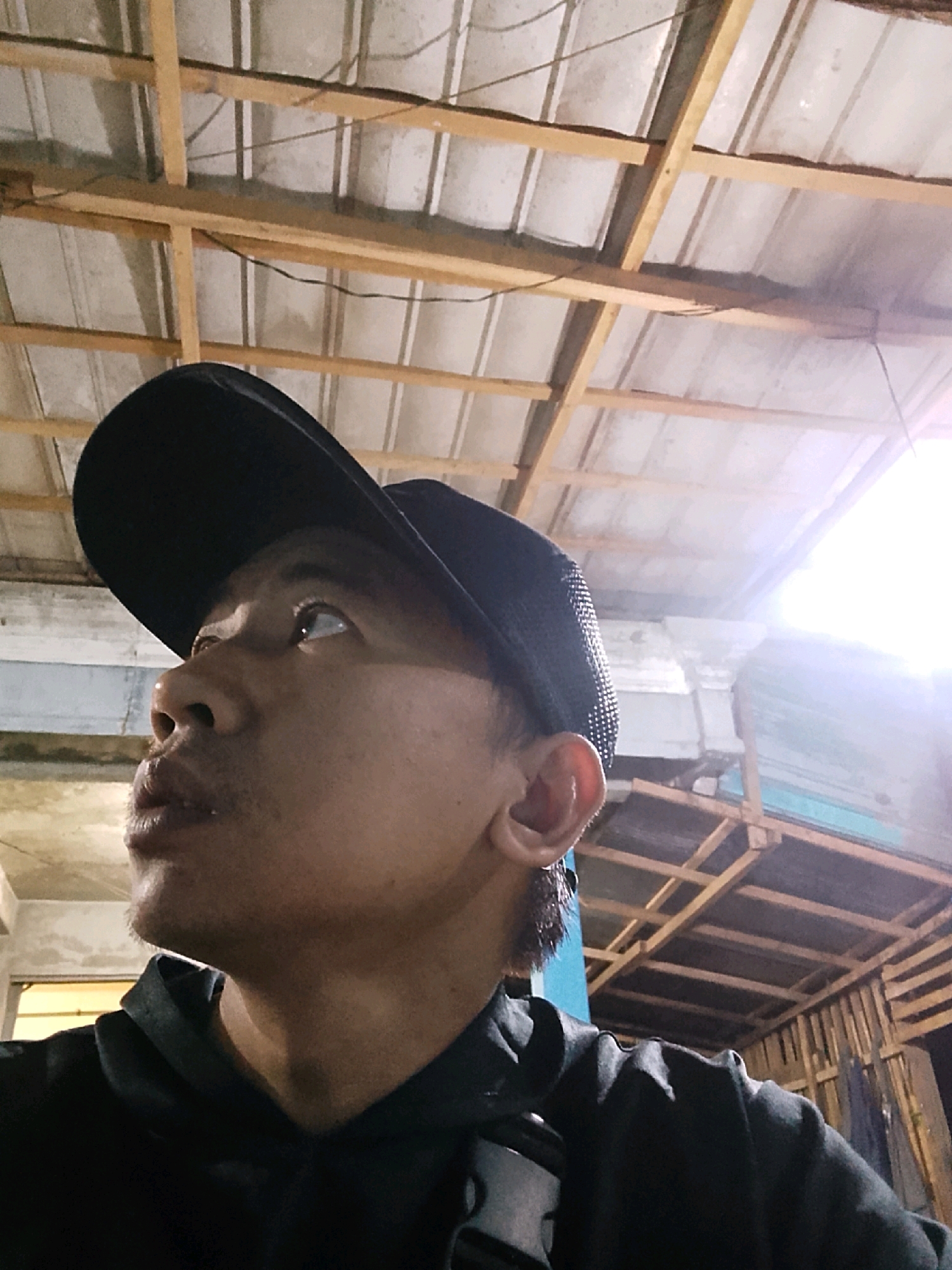 A close-up, low-angle, waist-up shot shows a person wearing a black baseball cap and a black hoodie. The person is looking upwards and to the right, with their mouth slightly open. The background reveals an unfinished ceiling with exposed wooden beams and corrugated metal sheeting. There is bright light emanating from the upper right of the frame. The scene appears to be an indoor or covered outdoor space, potentially under construction or in a rustic setting, possibly in Pelaihari, Indonesia, given the context.