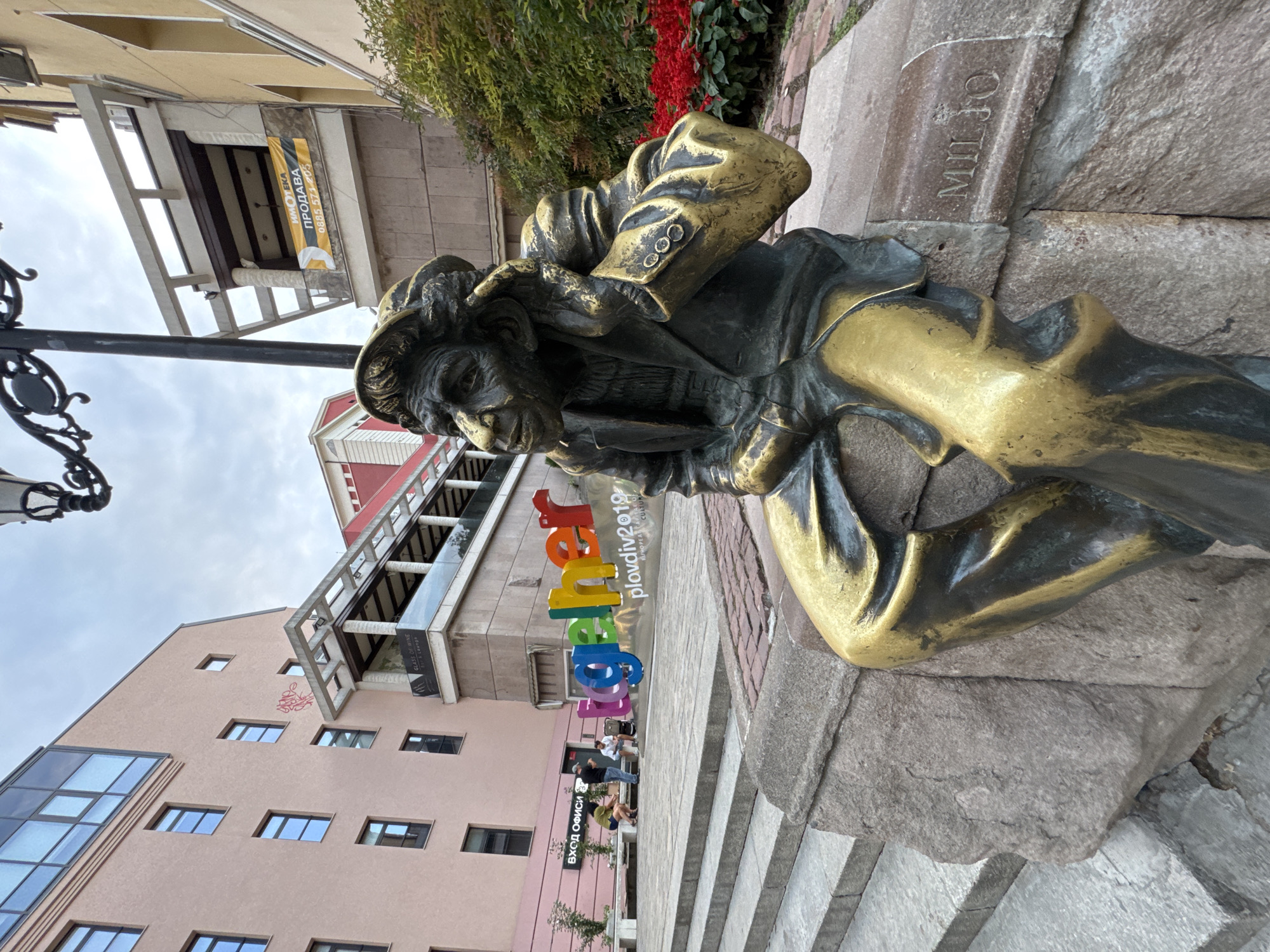 In Plovdiv, Bulgaria, a bronze statue of a man sits on a stone pedestal. The statue depicts a weathered man with a cap, wearing a suit and holding his hand to his ear as if listening. His suit is rendered with gold accents, creating a striking contrast with the darker bronze. The man's facial features are detailed, conveying an expression of contemplation or attentiveness.
Behind the statue, a vibrant, large-scale art installation spells out the word "together" in colorful, three-dimensional letters. Below this, the text "PLOVDiV2019" and "EUROPEAN CAPITAL OF CULTURE" are visible, indicating the location and context of the artwork.
The setting appears to be an outdoor public space, possibly a plaza or pedestrian area. Steps are visible to the left of the statue, leading upwards. In the background, a light pink building with multiple windows and some graffiti can be seen. Further back, another building with yellow facade is partially visible, with a banner that reads "ПРОДАВА" (FOR SALE) in Cyrillic. Red flowers are planted in a garden bed to the right of the statue, adding a touch of color to the scene. Several people are seated on a bench in the middle ground, engaged in conversation. The sky is overcast.