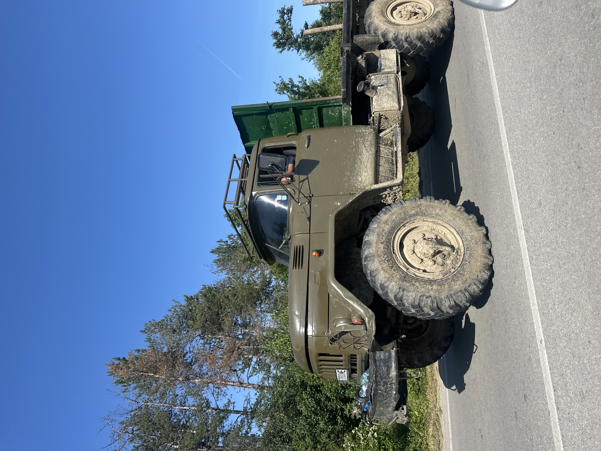 The image captures a large, olive-green truck with oversized tires driving on a paved road. The truck is heavily soiled with mud, indicating it has recently been off-road. It features a flatbed cargo area at the rear and a rack on the roof. A driver is visible inside the cab. The background consists of trees and a clear blue sky. The scene appears to be in a rural or semi-rural setting, possibly near Prisovo, Bulgaria, given the context provided. The overall impression is one of a rugged, utility vehicle in use.