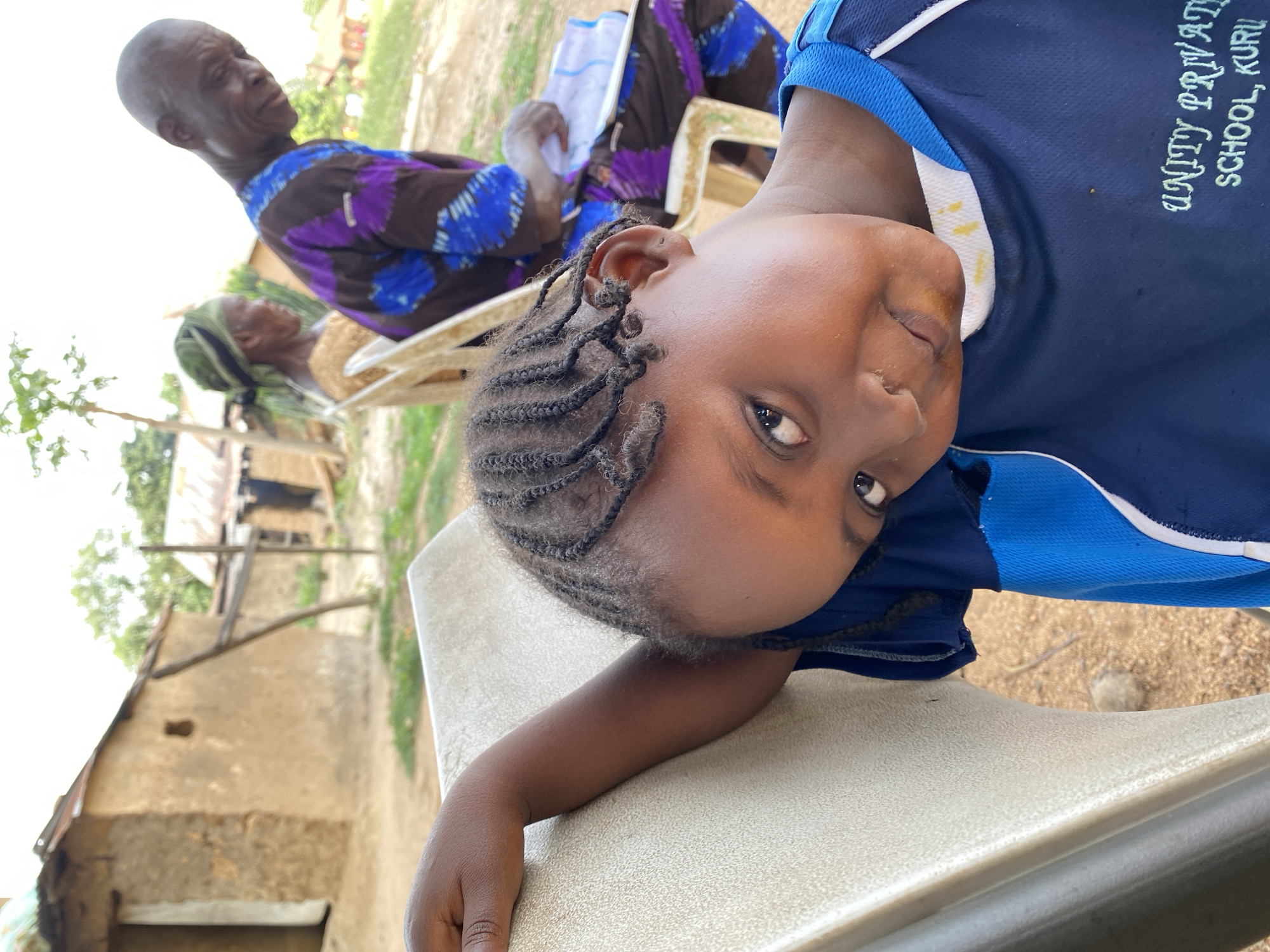 A young girl wearing a blue school uniform sits on a white plastic chair. "Unity Private School, Kuru" is visible on her shirt. Her hair is styled in braids. Behind her, an elderly man in a colorful patterned shirt sits with paperwork. An elderly woman with a head covering also sits in a chair. The backdrop shows a simple dwelling, suggesting a rural environment in Kuru Dungus, Nigeria.