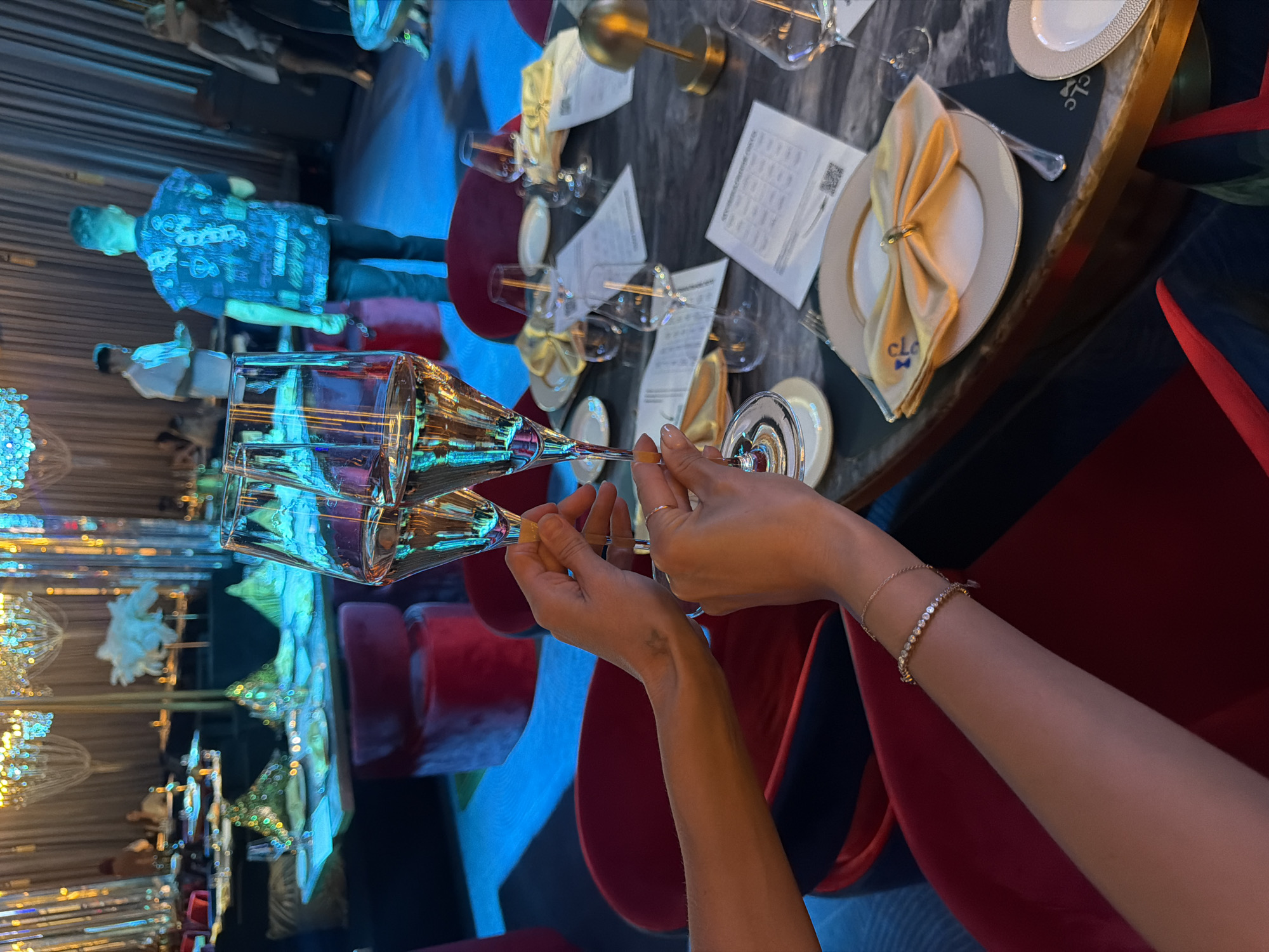 The image shows an indoor dining scene, possibly in a high-end restaurant or event venue, potentially in Choeng Thale, Thailand. The camera is at eye-level, pointed at a dining table setting.
In the foreground, two glasses, likely wine glasses, are held up by a person. The glasses are partially filled with a clear liquid. The person's hands are visible, with manicured nails painted with a yellow hue. A delicate bracelet adorns one wrist.
The table is set with plates, napkins, silverware, and menus. A gold-colored napkin ring and a "CLG" monogram on the napkin are visible. Additional glassware suggests a formal dining experience.
The background features other diners and staff. There is a blue lighting effect. Ornate chandeliers can be seen. The overall atmosphere appears luxurious. Red chairs around the table provide a contrast to the cooler tones of the scene.