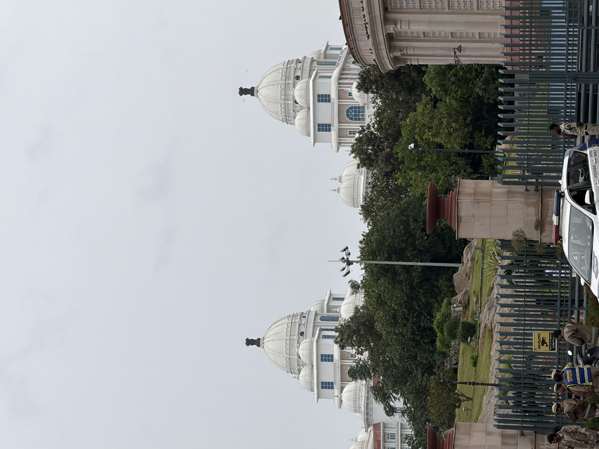 The image shows the impressive architecture of the Telangana High Court in Hyderabad, India, under a cloudy sky. The building features multiple white domes, the largest of which is topped with the Ashoka Chakra. Several smaller domes surround the main dome, and the structure has numerous windows. In the foreground, a green metal fence encloses the grounds, beyond which is a landscaped area with rocks and trees. Several security personnel are visible near the fence and a police patrol car. A yellow sign with Telugu text and "CCTV" is also mounted on the fence.