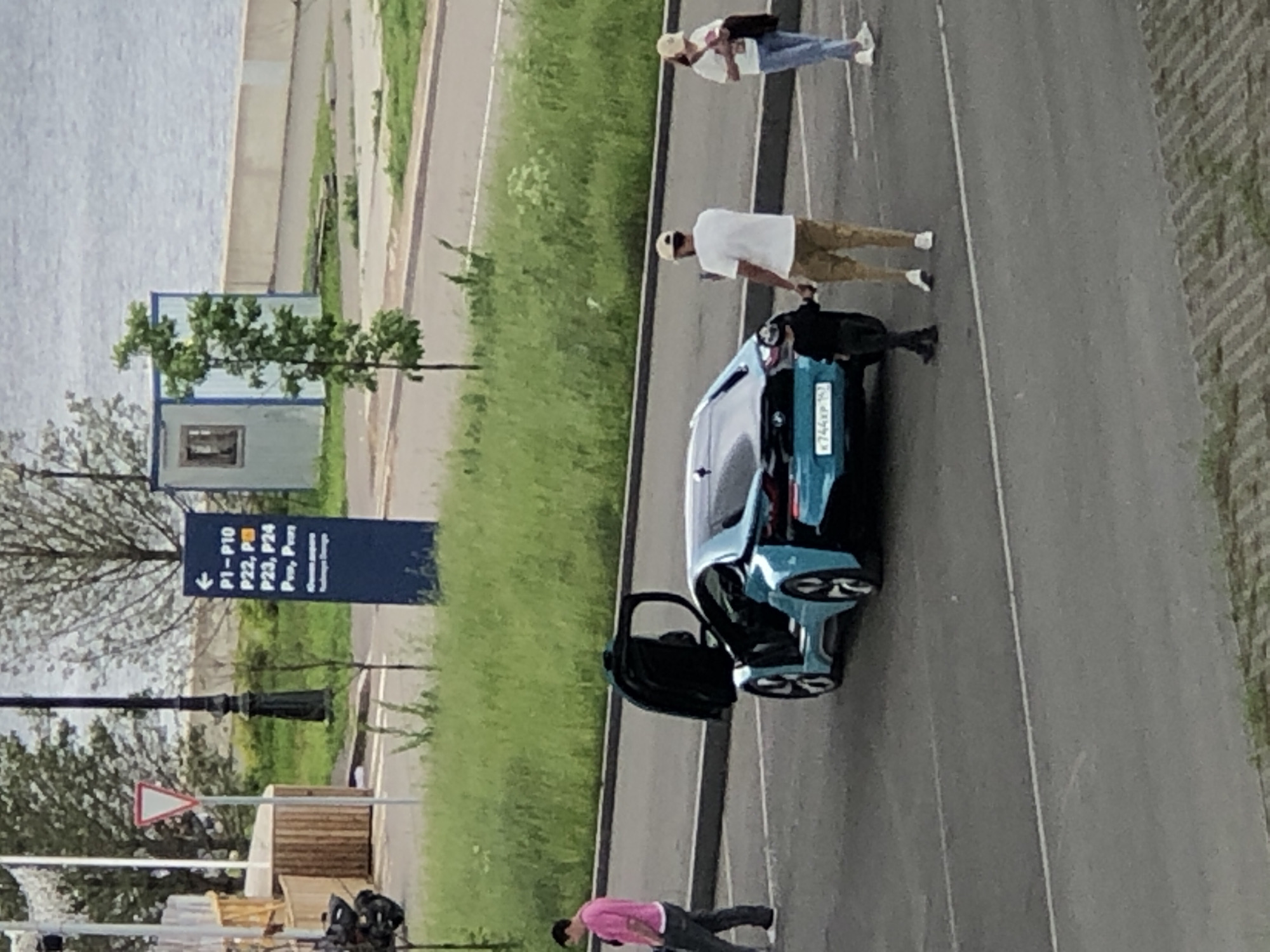 In an outdoor, paved area resembling a parking lot, a bright blue sports car with its dihedral doors open is the central focus. The car is positioned at an angle, facing slightly towards the right of the frame. To the left of the car, a person in a pink shirt and dark pants is walking away from the viewer. To the right of the car, a man wearing a white t-shirt and tan pants, holding a phone in his right hand, stands with a young child. The child, dressed in dark clothing, is holding the man's left hand. Further to the right, a woman in a white t-shirt, blue jeans, and a light-colored cap is walking. In the background, there is lush green grass and trees. A dark blue signpost with white text and arrows is visible on the left side of the frame, indicating parking zones. There is also a light blue temporary building with a small window to the right, near a body of water. The overall scene suggests a casual outing or visit to an area with amenities.