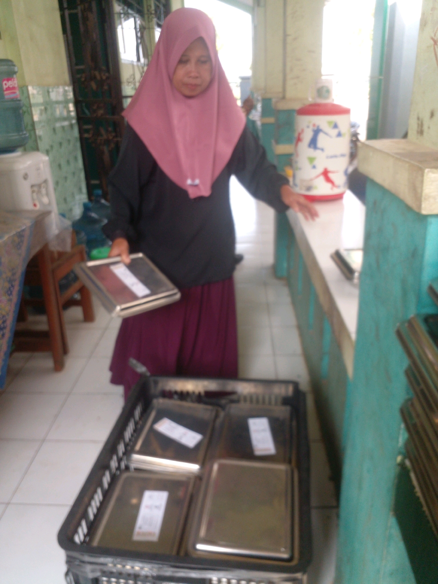 A woman wearing a pink hijab and a black long-sleeved shirt stands in what appears to be a dining area, possibly a school or communal canteen, in Banyuputih, Indonesia. She is holding a silver tray containing food items, and there is a black plastic crate filled with similar trays on the floor in front of her. Behind her, a water dispenser is visible, along with a decorative thermos. The environment suggests a practical, perhaps institutional setting, with tiled floors and utilitarian furniture.