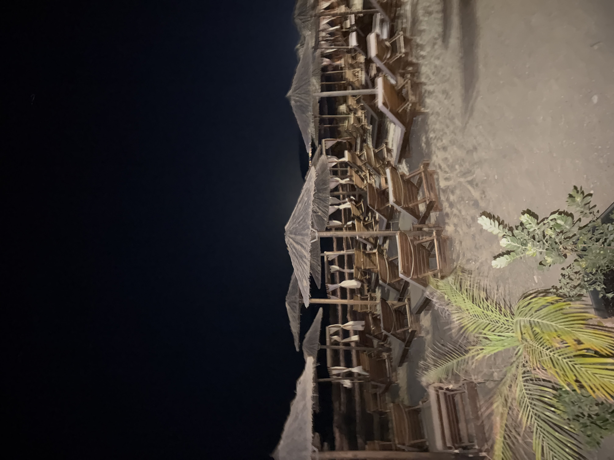An outdoor scene at night on a sandy beach in Georgioupoli, Greece, features rows of empty wooden lounge chairs and thatched umbrellas set up on the sand. The sky above is dark. In the foreground, to the left, a palm frond plant is visible. The lighting in the scene suggests it is artificial.